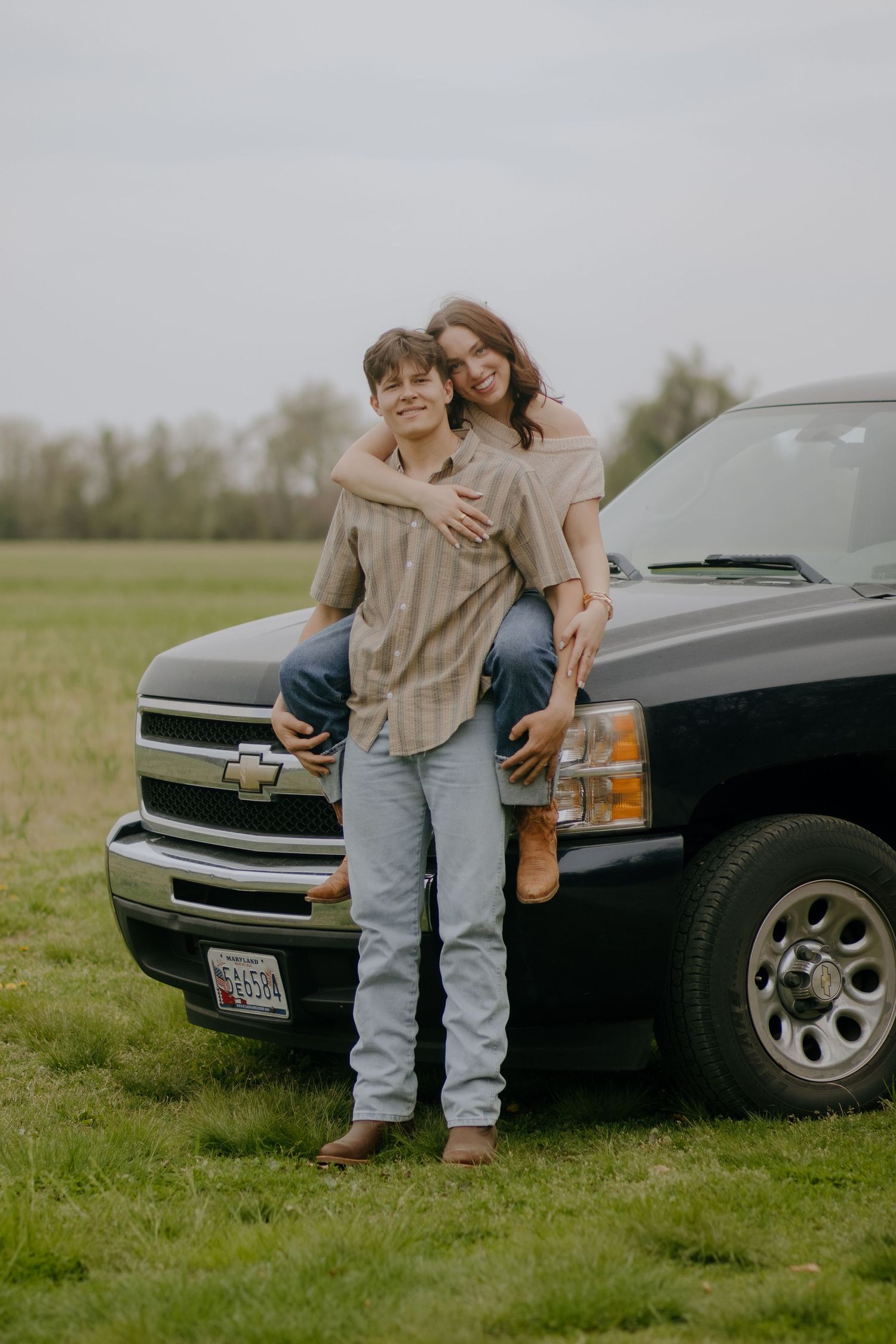 A couple poses affectionately next to a black Chevrolet truck in a grassy field.