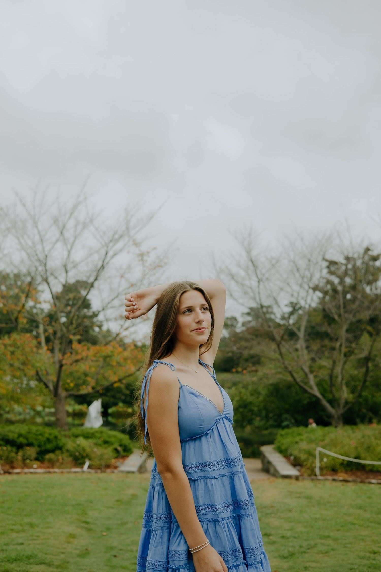 A woman in a blue denim dress stands in a garden setting on an overcast day.