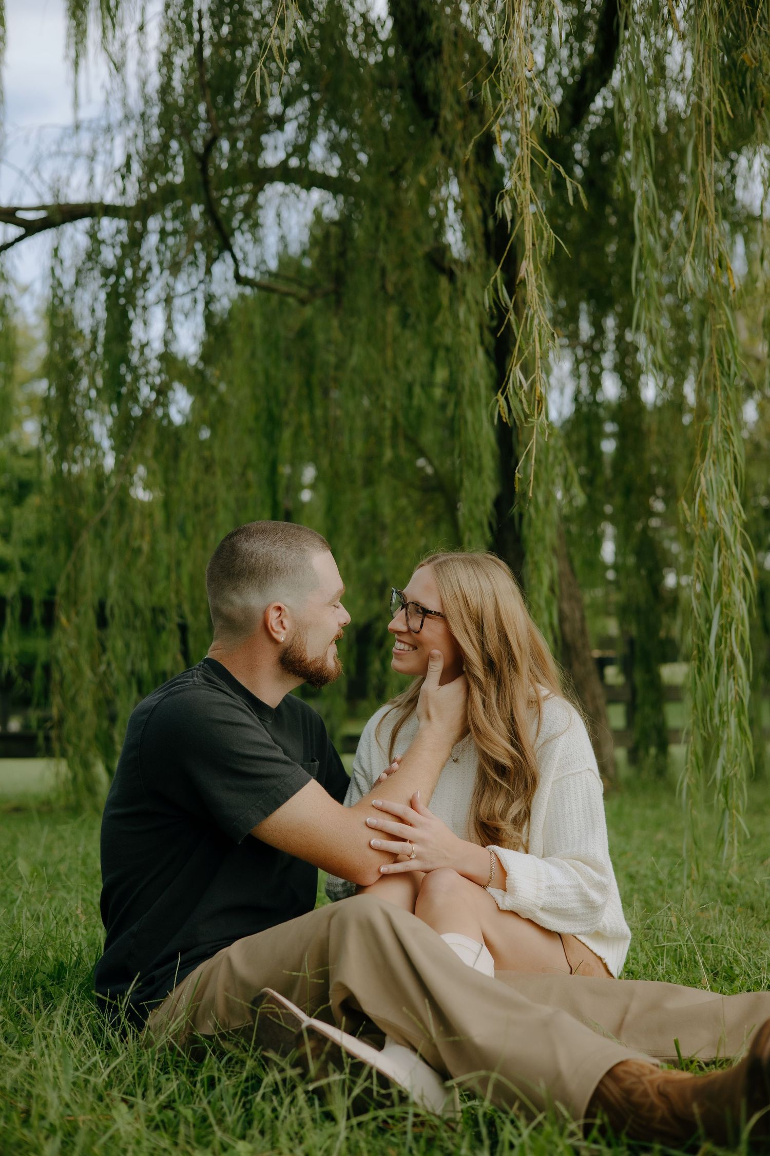 A couple shares a romantic moment while sitting beneath a weeping willow tree on a grassy lawn.