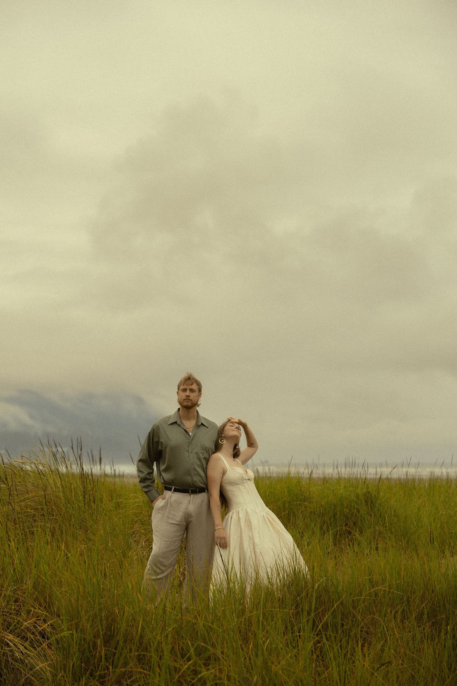 A couple stands in tall green grass under a cloudy sky, woman in white dress leaning on mans shoulder.