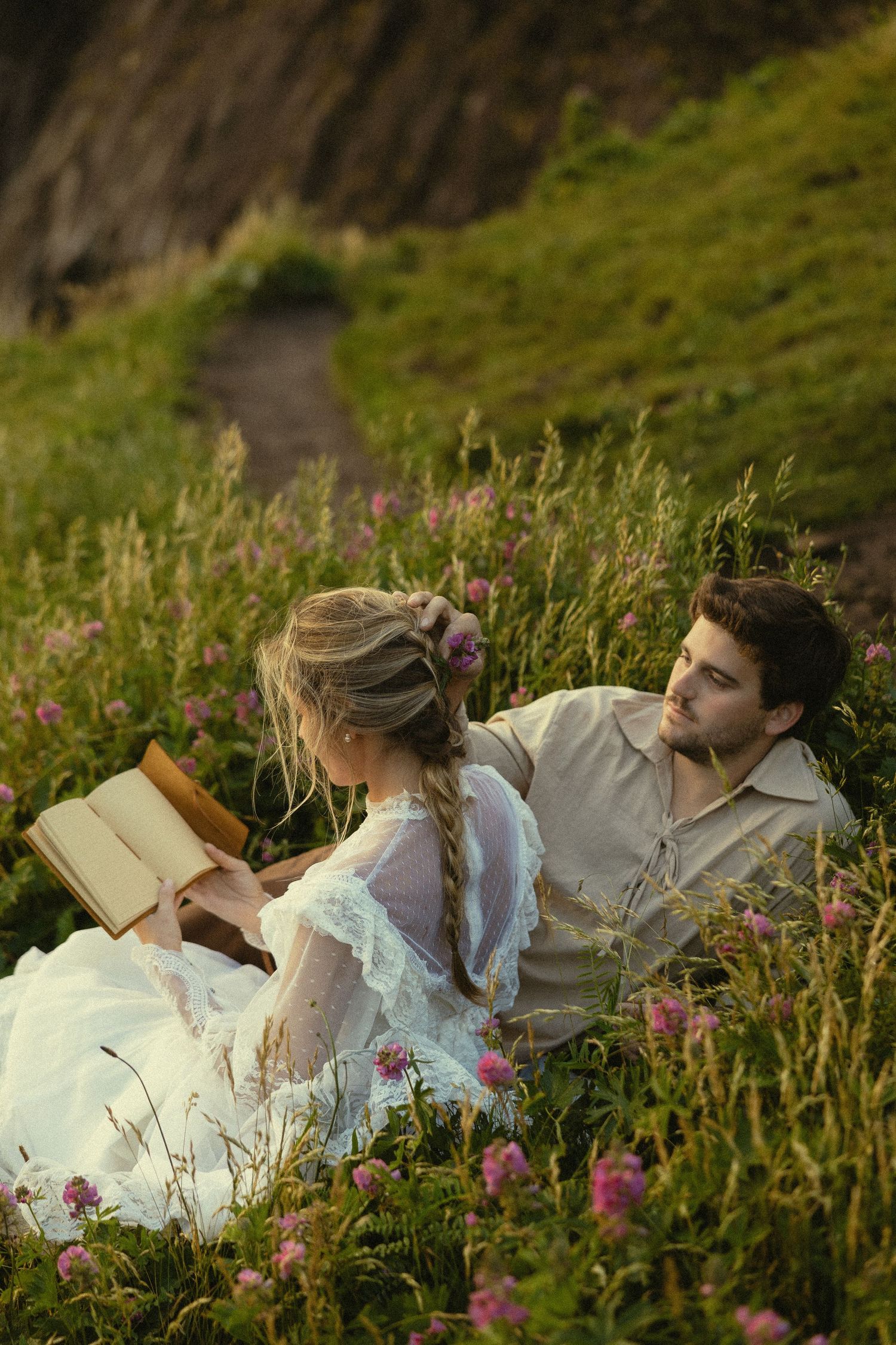 A couple relaxes in a wildflower meadow, woman in white lace dress reading a book while man gazes at her adoringly.
