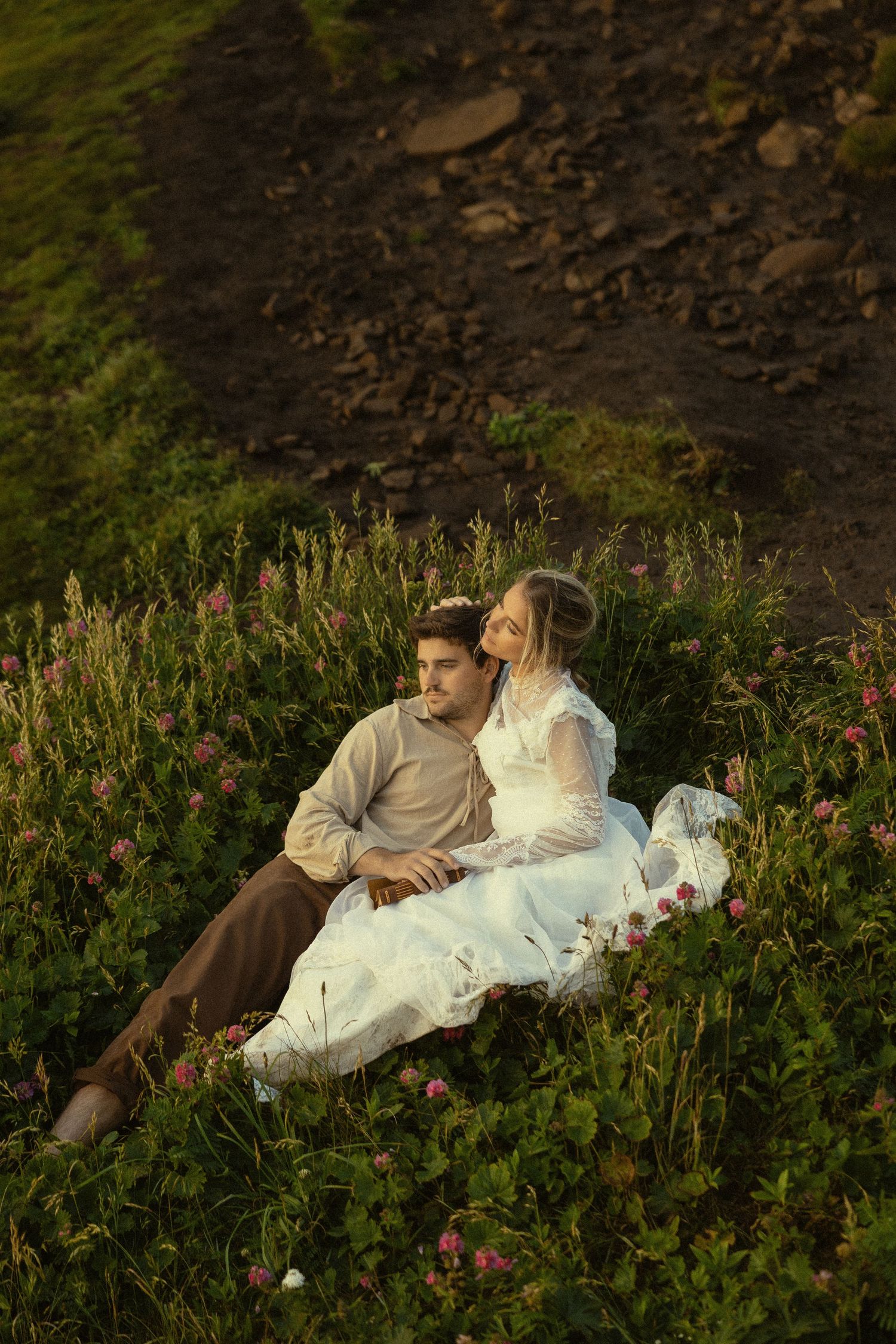A couple in romantic attire sits together in a wildflower meadow, the bride in white lace leaning on her groom.
