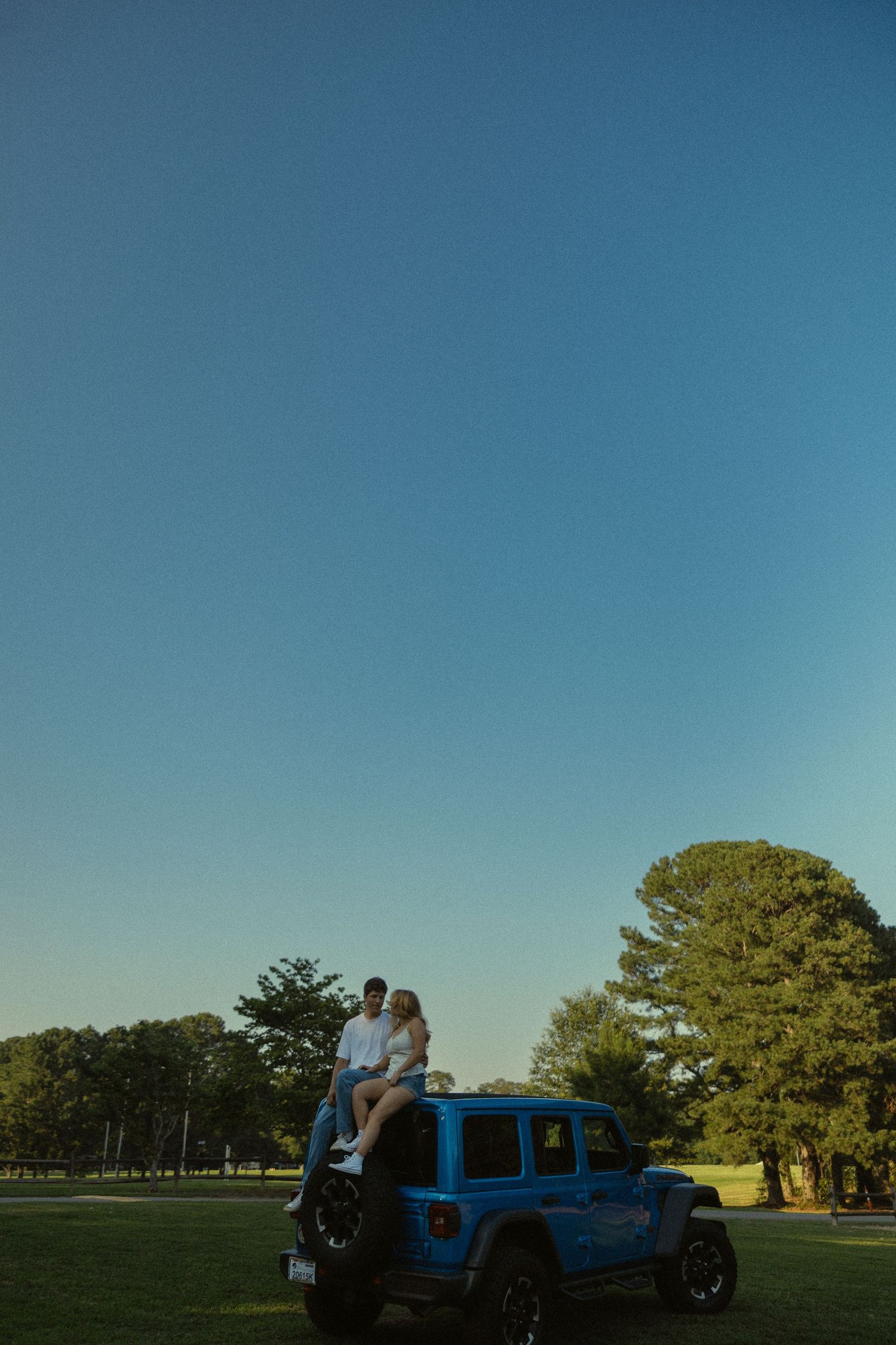 A couple sits atop a blue Jeep Wrangler on a sunny day with clear blue skies and green trees.