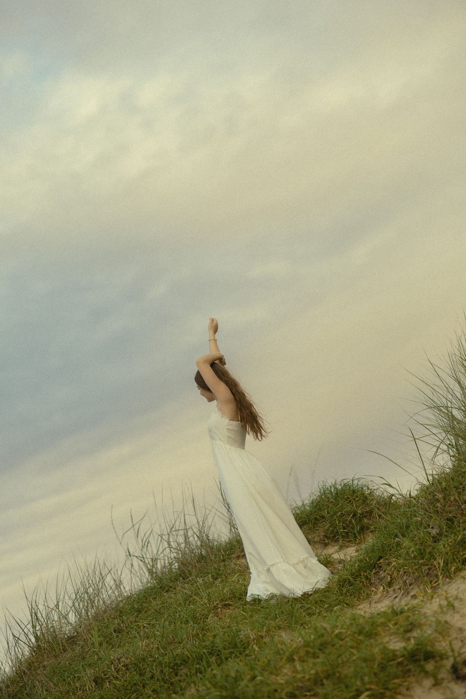 Woman in white dress stands on grassy dune with arms raised toward cloudy sky at dusk.