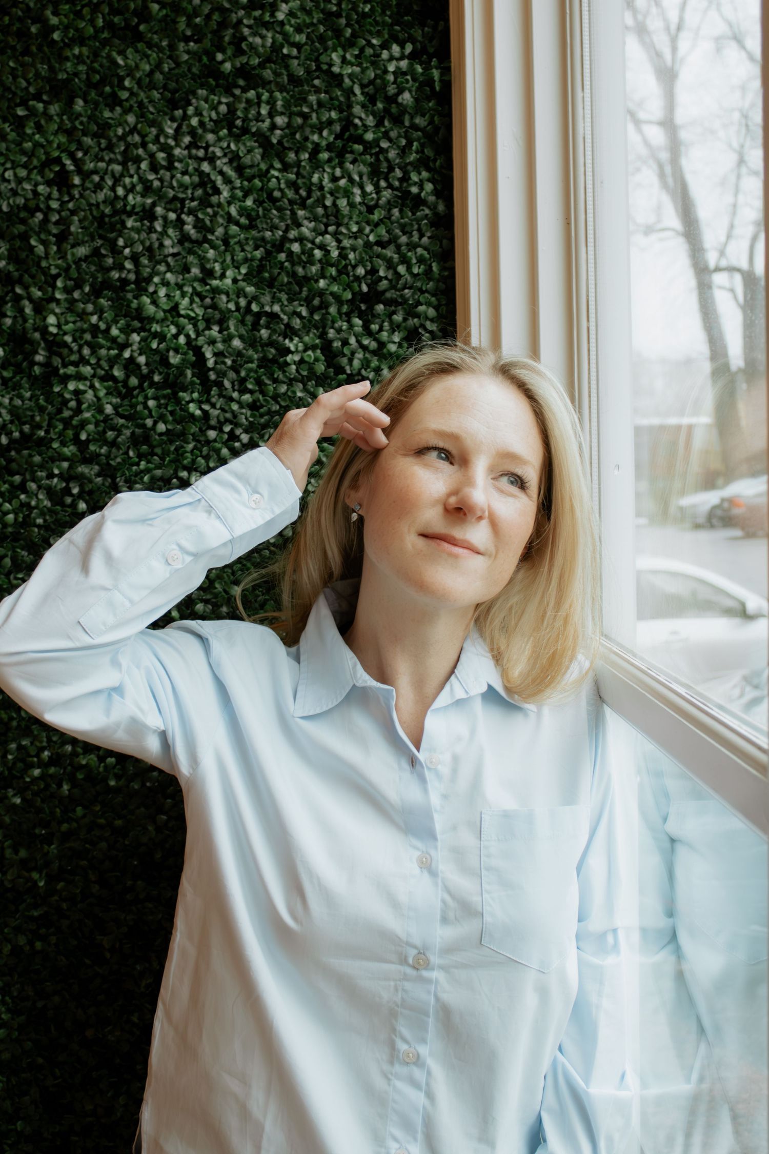 Person in light blue button-down shirt posing by window with greenery backdrop.