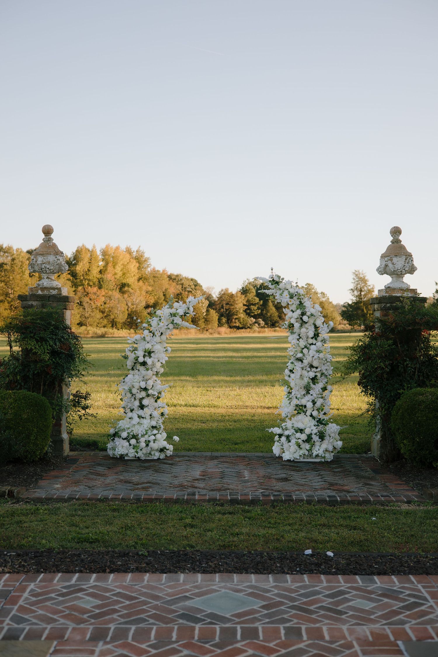 White floral wedding arch arrangements on brick pathway at sunset.
