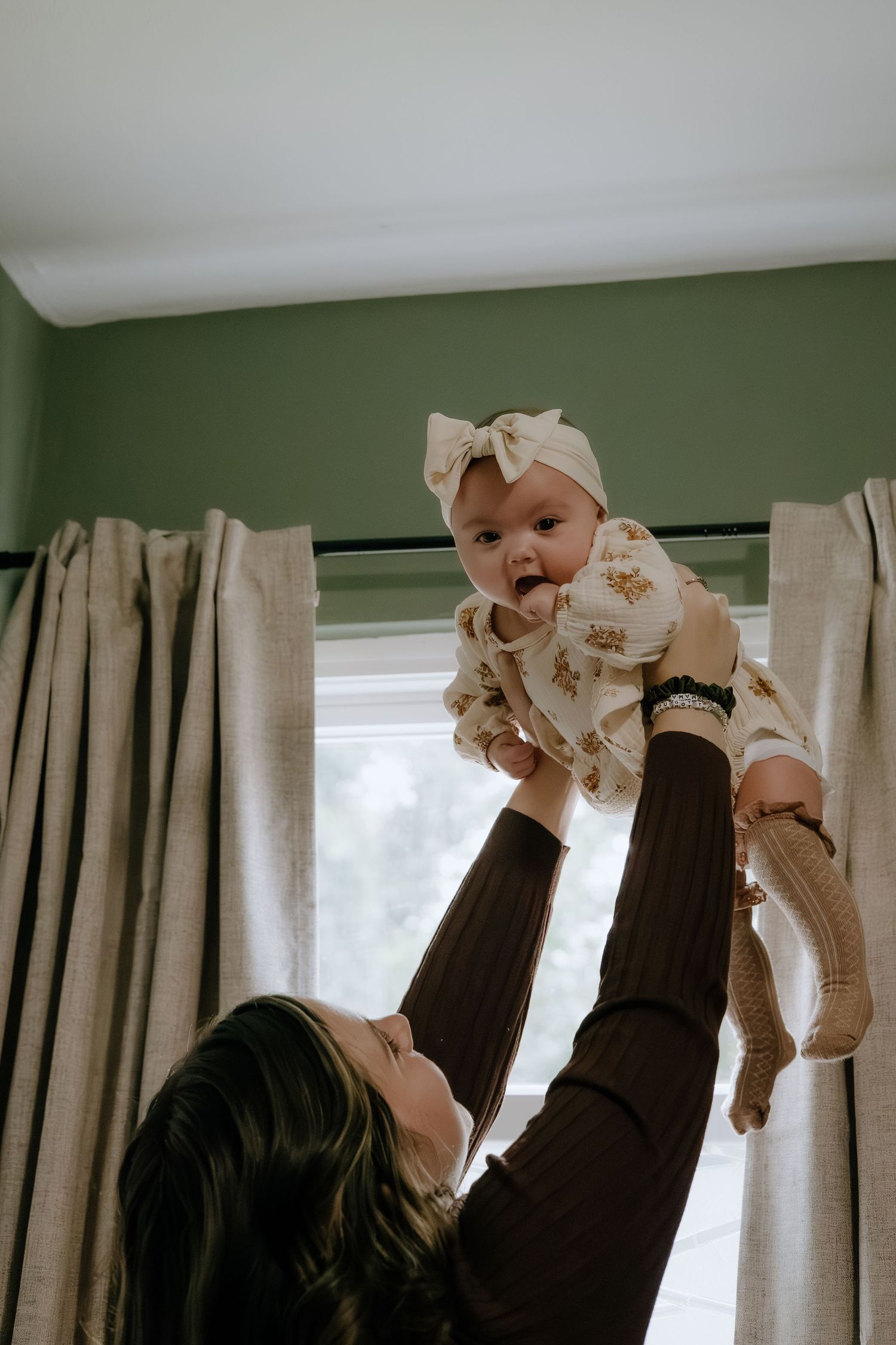 Baby wearing a cream dress and bow being lifted high in a living room with green walls.