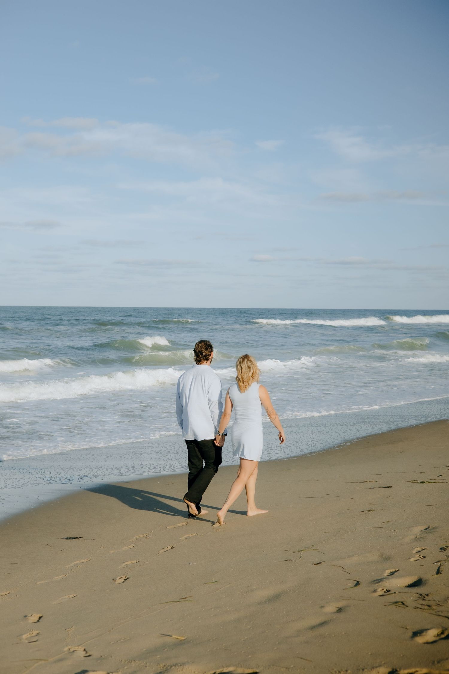 Couple holding hands walking barefoot on sandy beach with ocean waves, wearing casual light blue outfits on a sunny day.