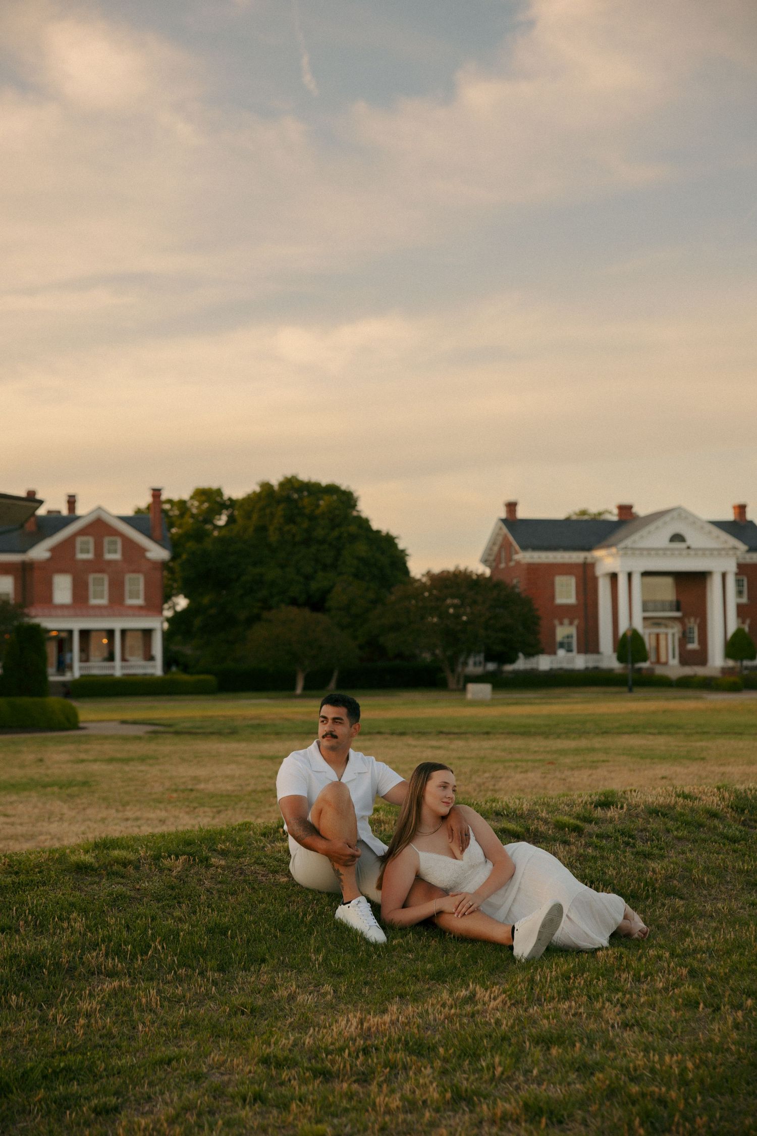 Couple sitting together on green grass at sunset, with historic red brick buildings and trees in the background.