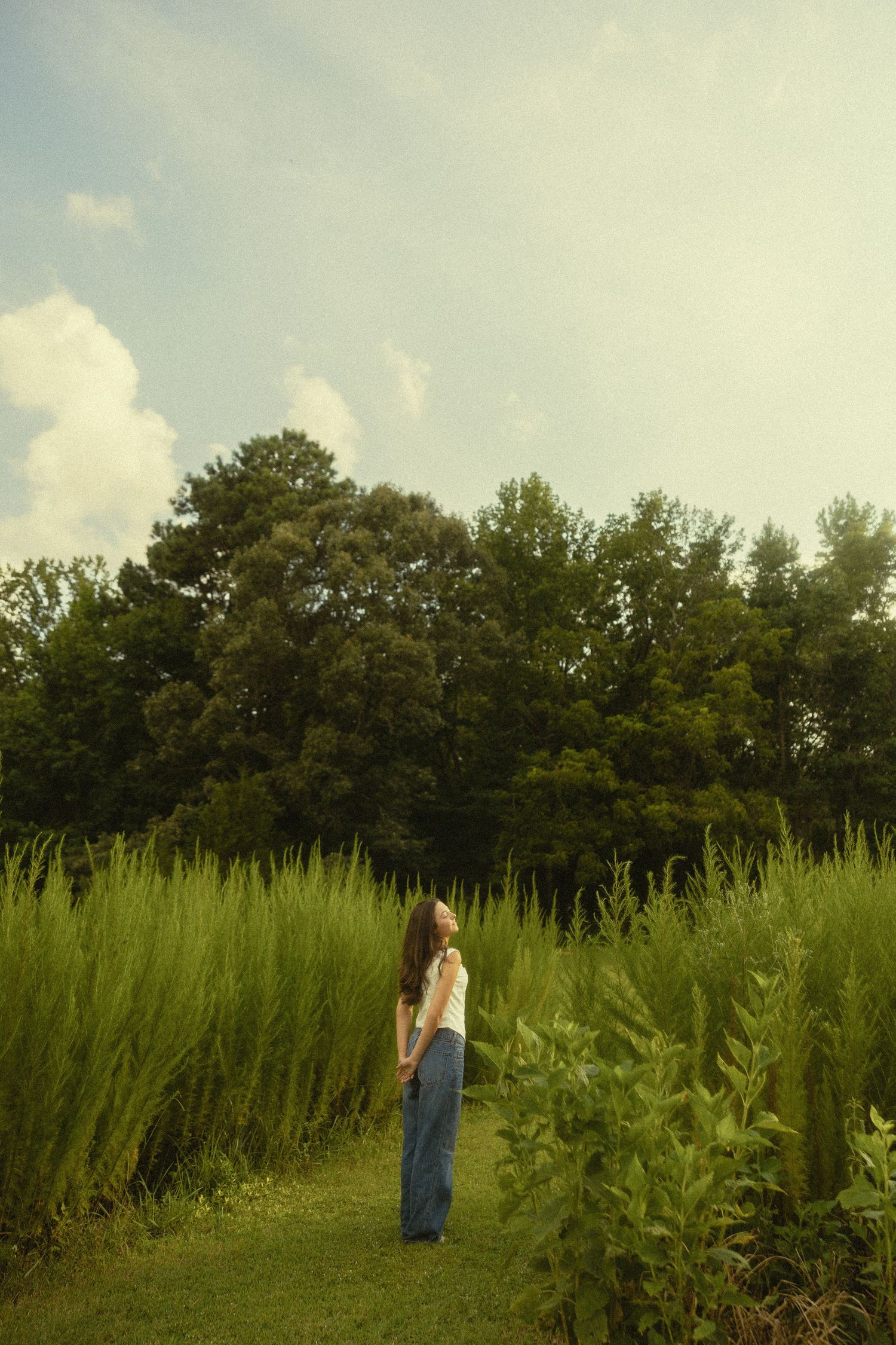 A young woman in white top and blue jeans stands in a lush green field surrounded by tall grass and trees.