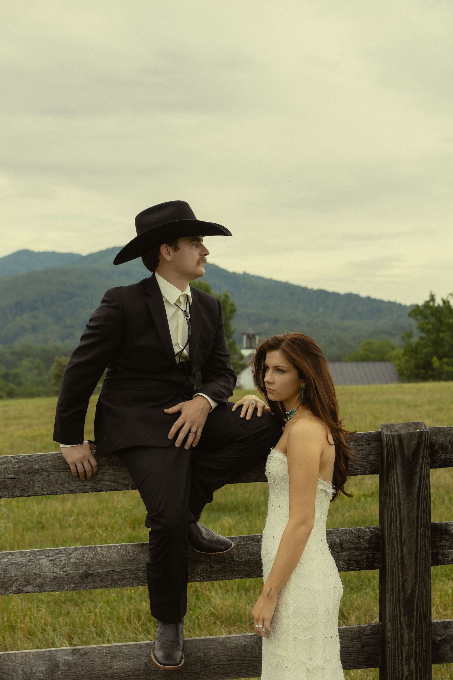 A bride in a white lace gown and groom in a black suit with cowboy hat pose by a rustic fence with mountain views.