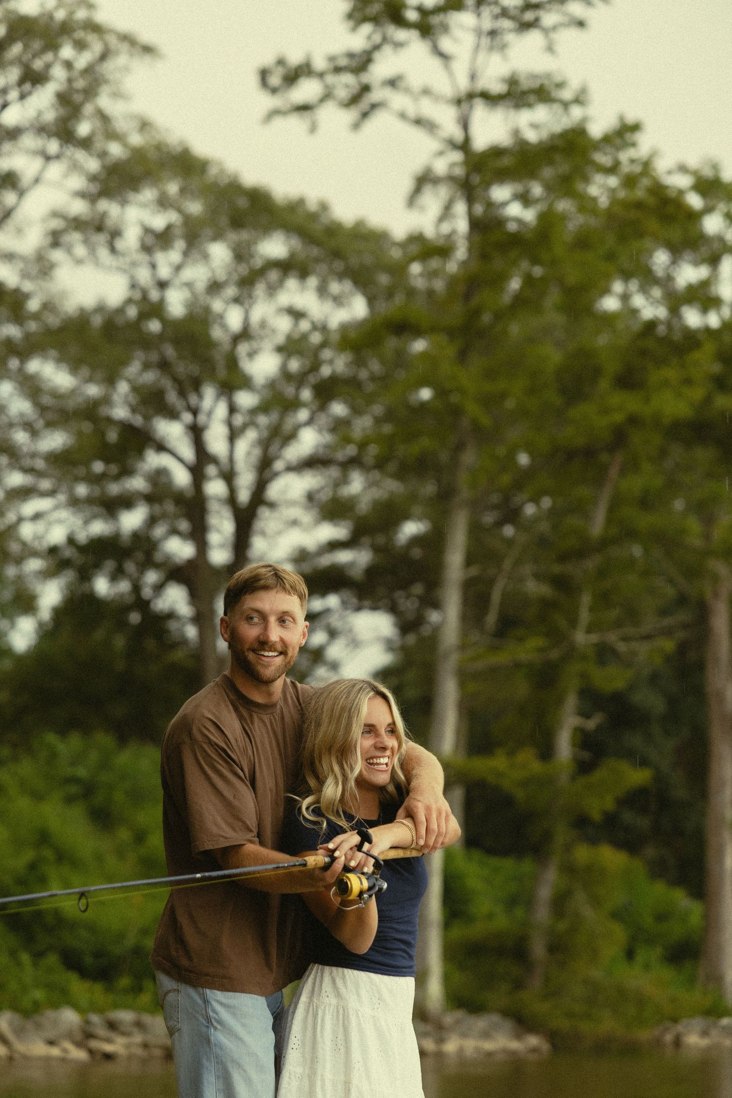 Happy couple smiling while fishing together by a lake surrounded by lush green trees outdoors.