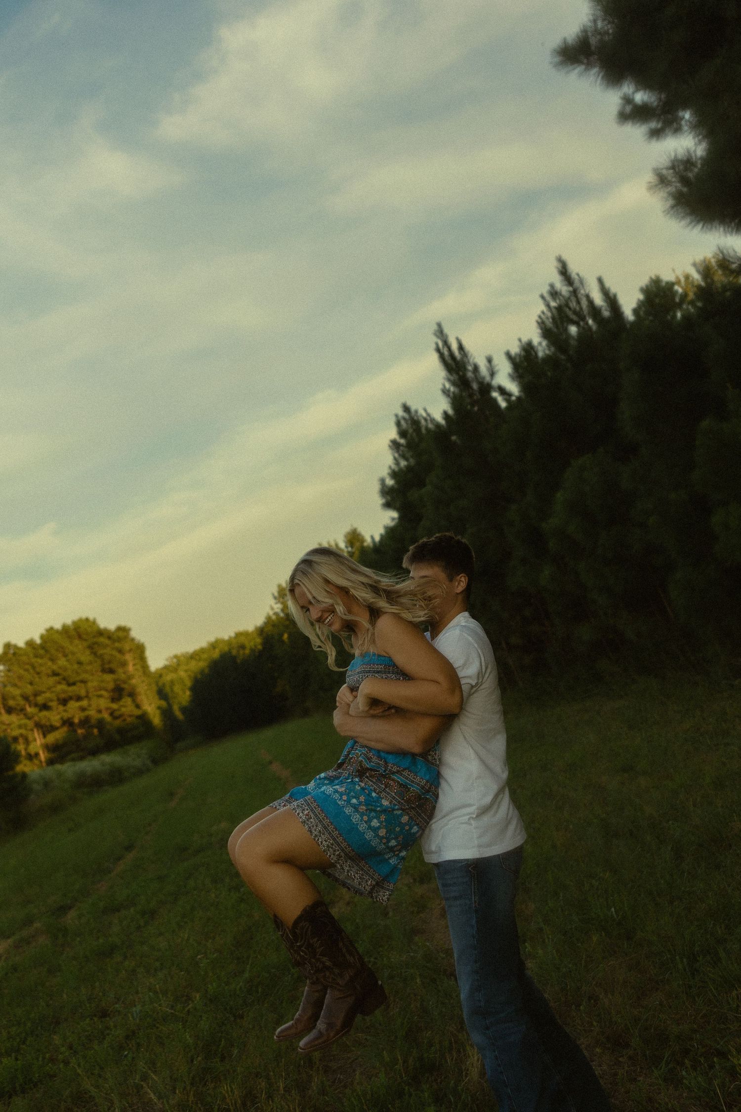 Couple dancing and embracing in a green field at golden hour with trees in the background.