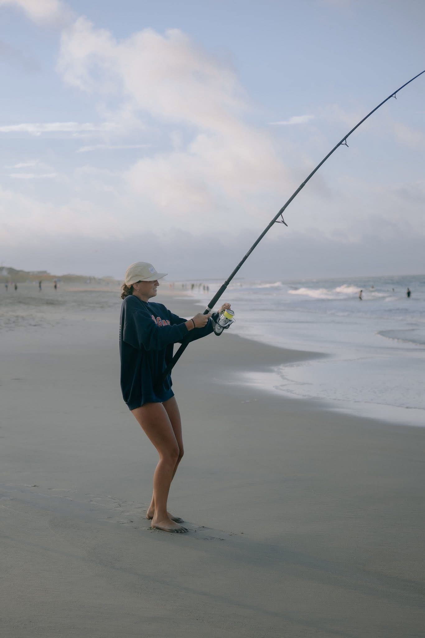 Woman surf fishing on a sandy beach, casting a long rod into ocean waves under a cloudy sky.