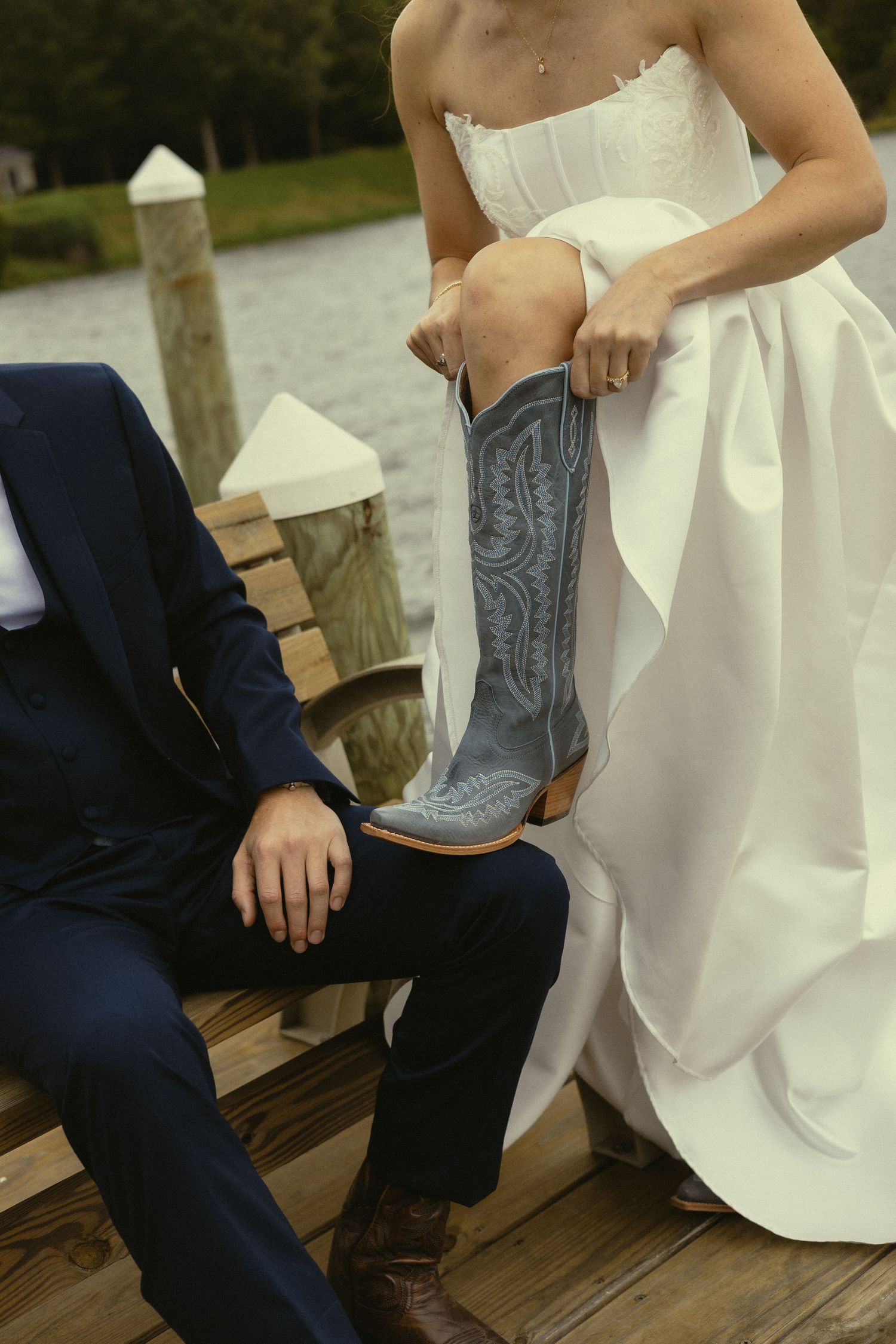 Bride in white strapless gown wearing blue cowboy boots on a wooden dock beside groom in navy suit.