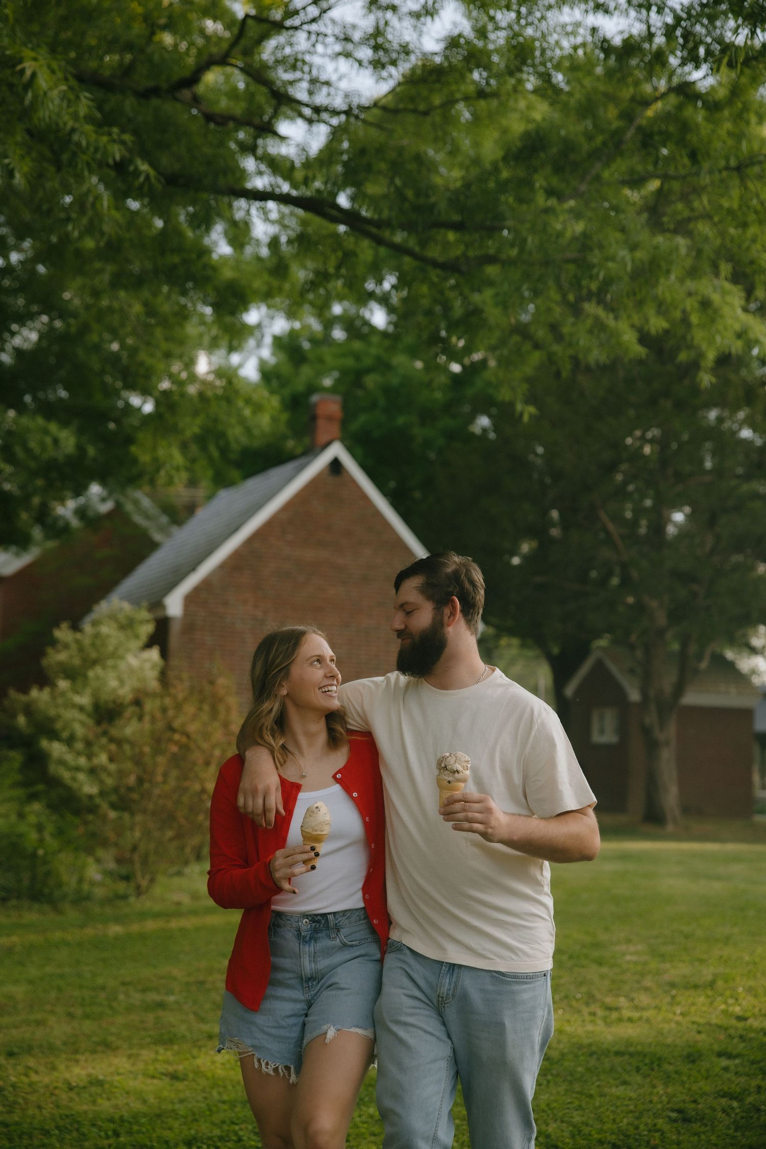 A smiling couple holds ice cream cones while standing together on a green lawn with a brick house and trees behind them.