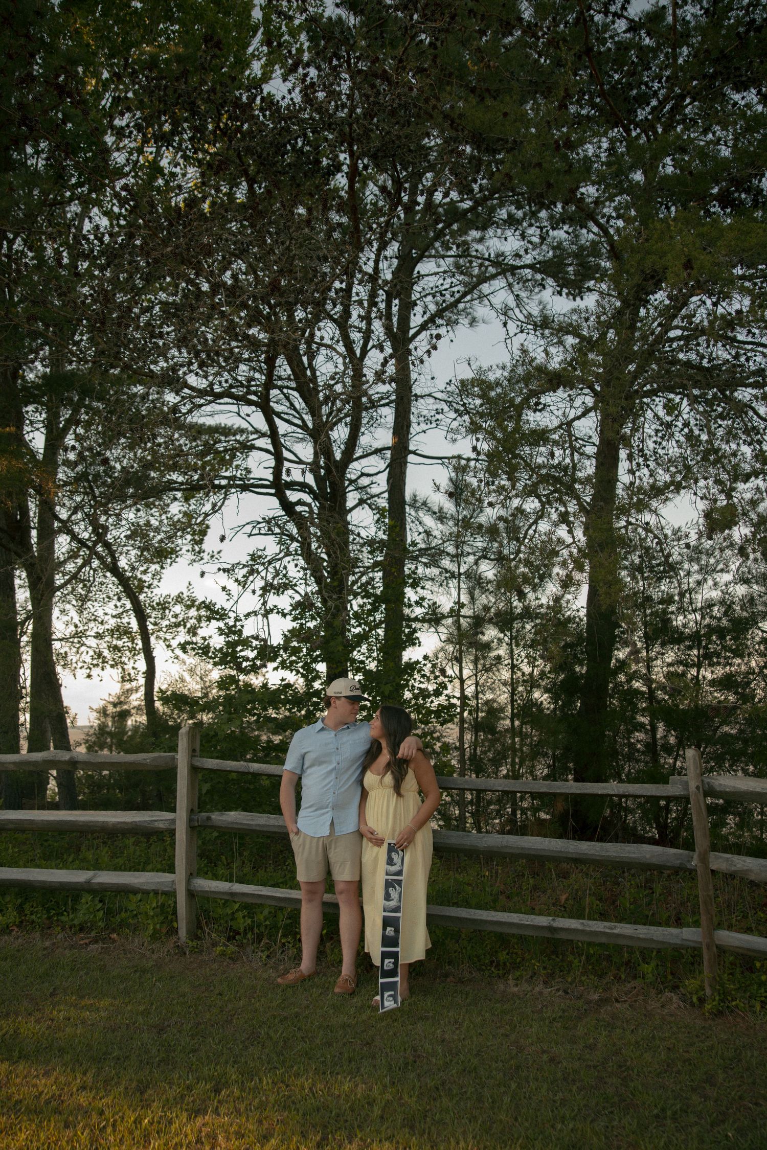 Couple standing by wooden fence at dusk among tall trees, woman in yellow dress holding ultrasound strip during pregnancy announcement.