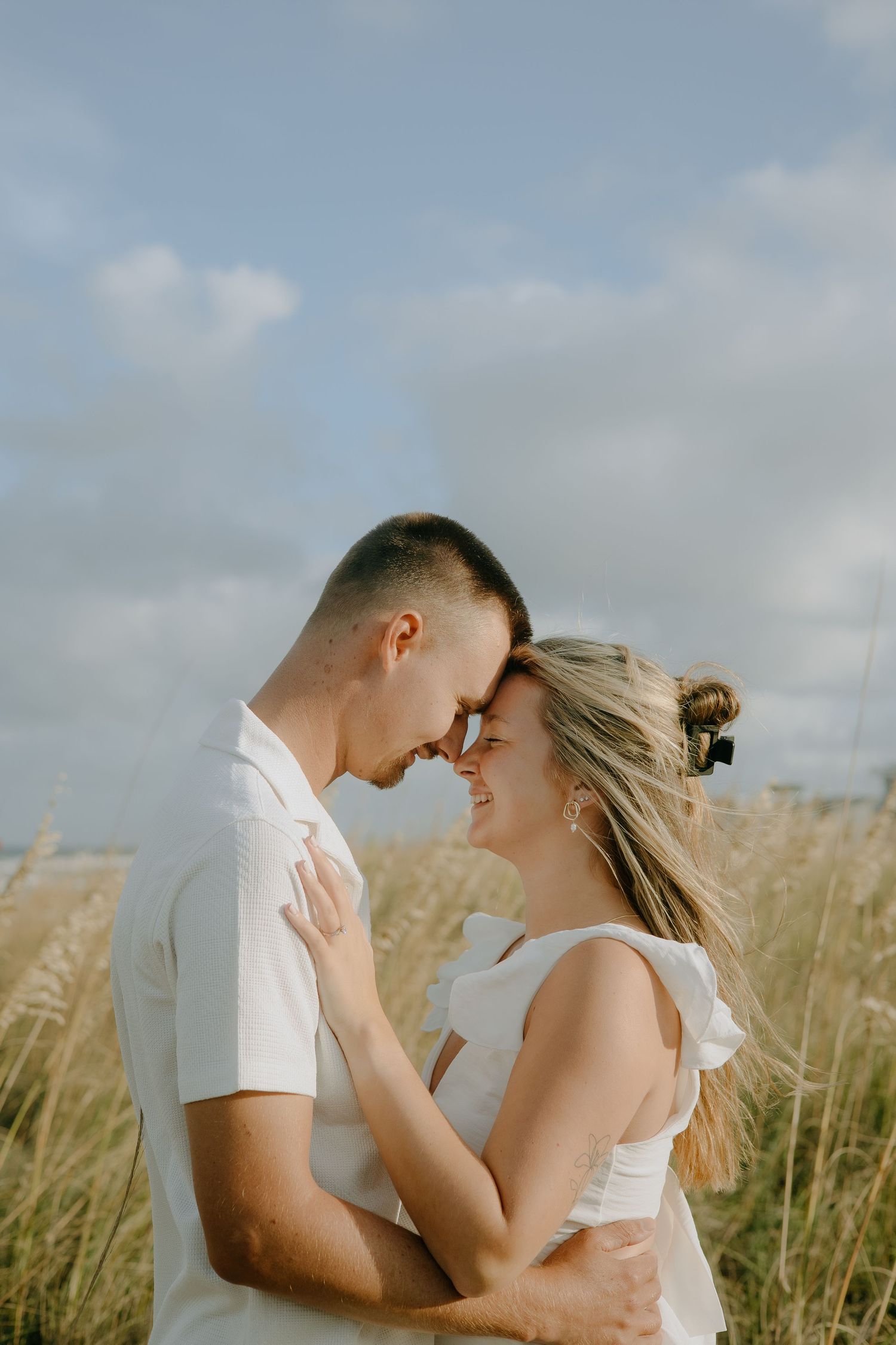 Couple in white outfits sharing a romantic moment in a golden field under a cloudy sky.