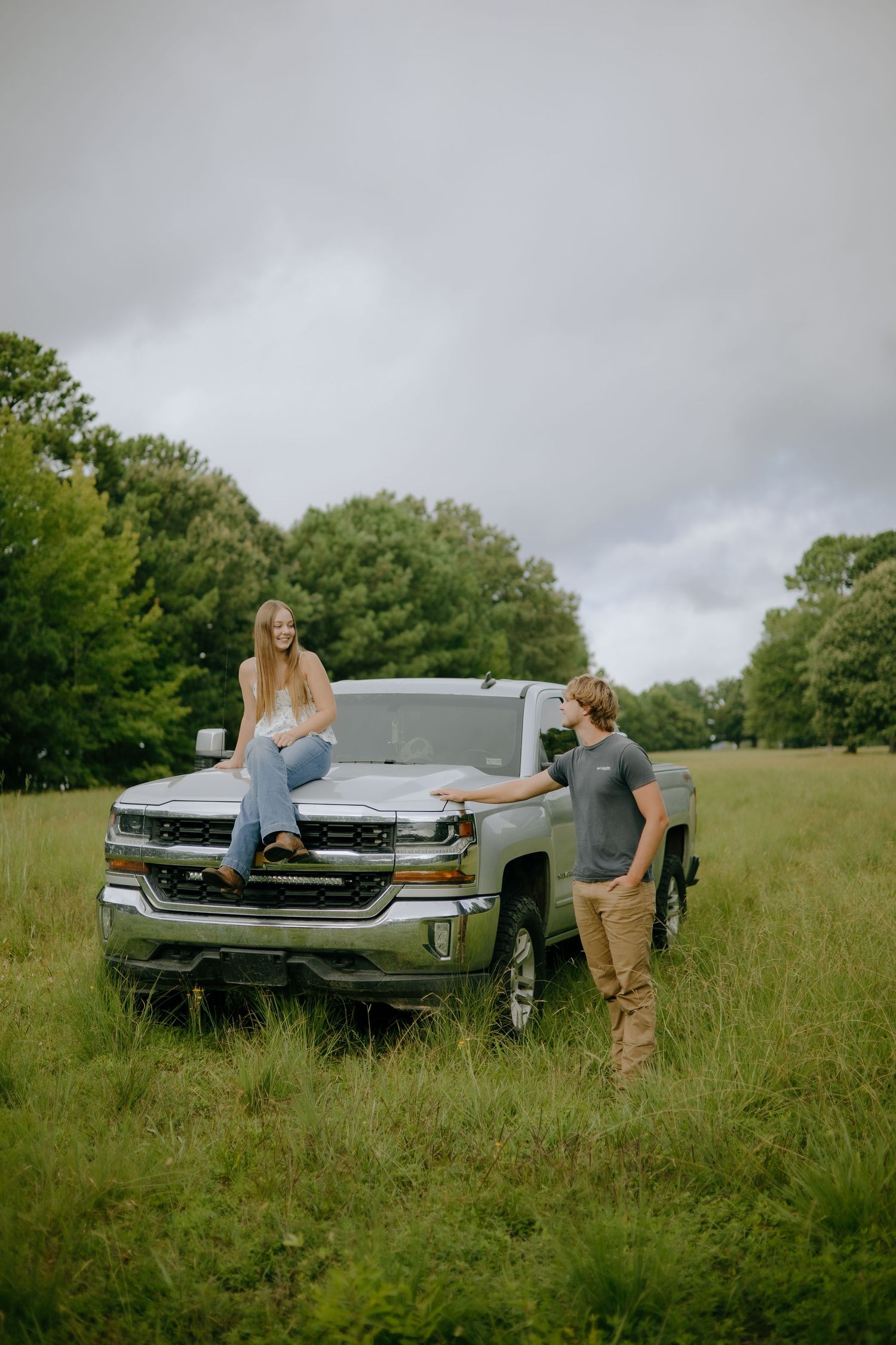 Friends hanging out around a silver Chevrolet Silverado pickup truck in a rural field setting.