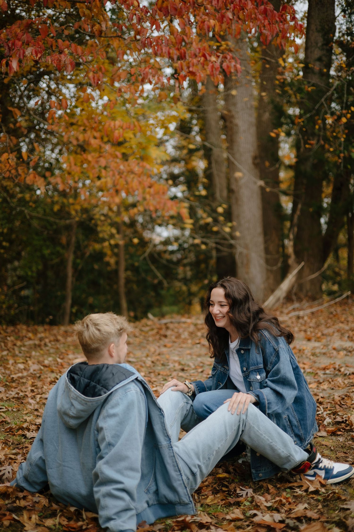 Two people in denim jackets laying on autumn leaves having a playful moment in the woods.