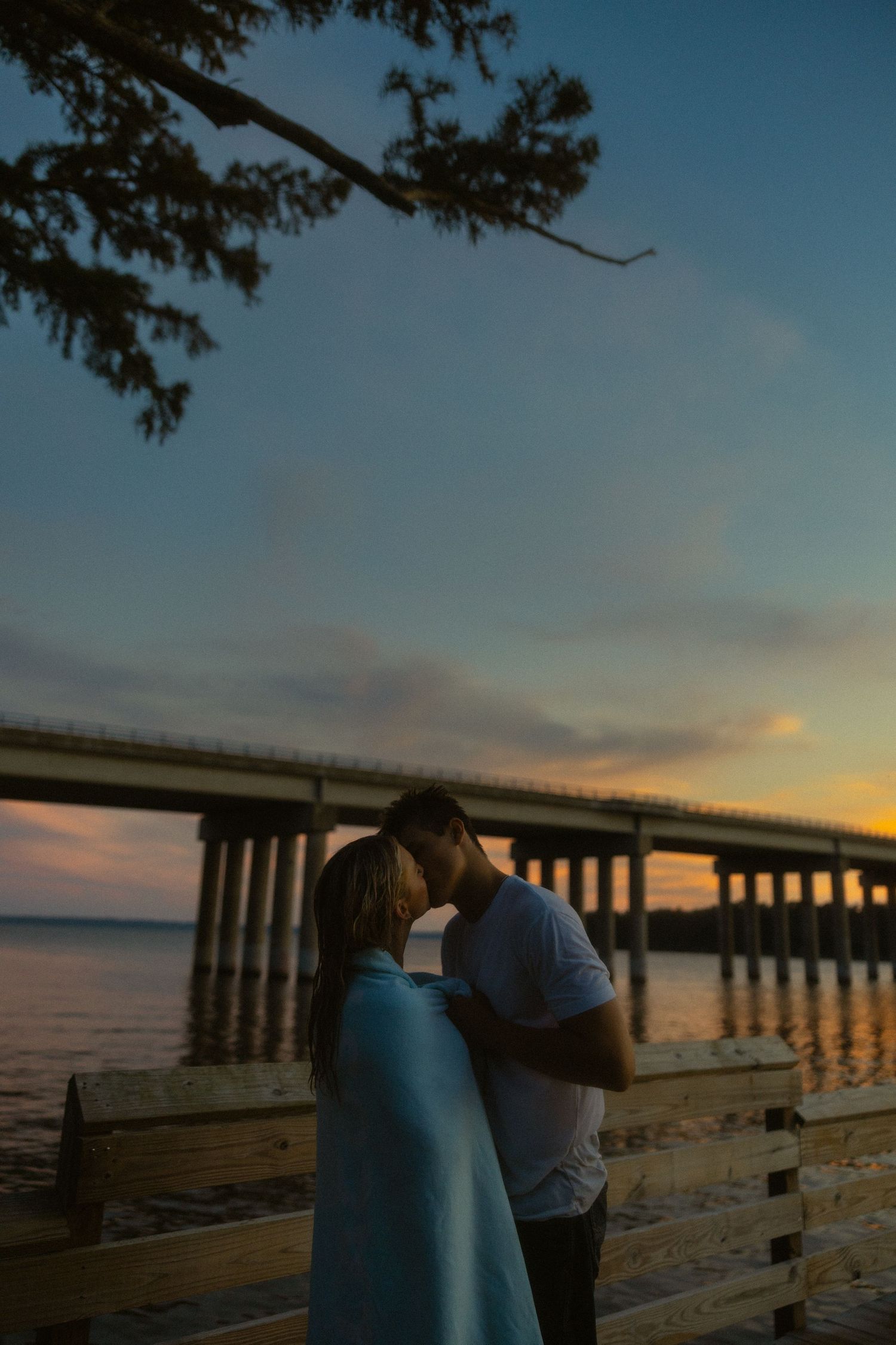 Couple wrapped in blue blanket sharing romantic moment at sunset near waterfront bridge at dusk.