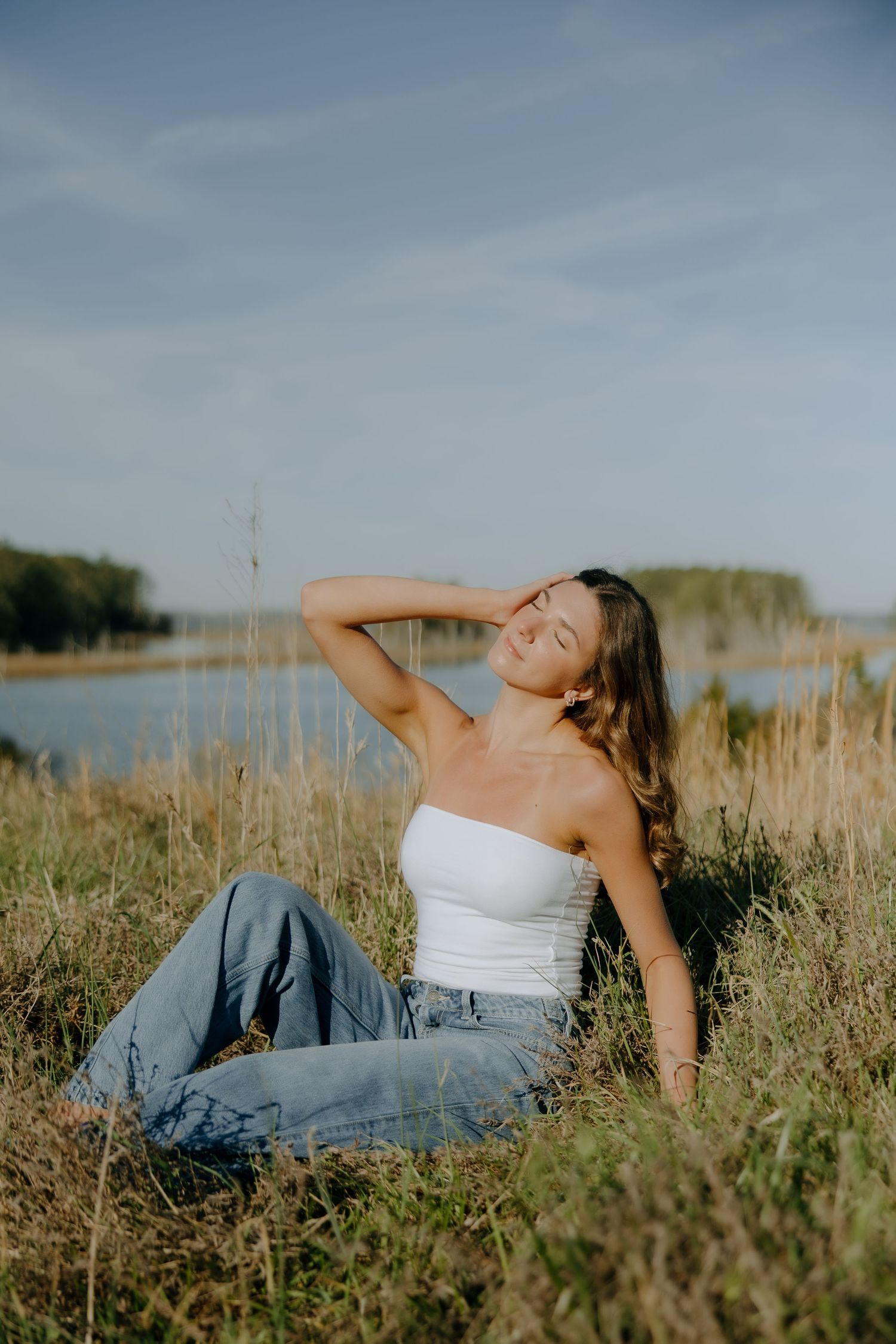 Young woman in white tube top and jeans sitting in golden grass by a lake, enjoying the sunshine with eyes closed.