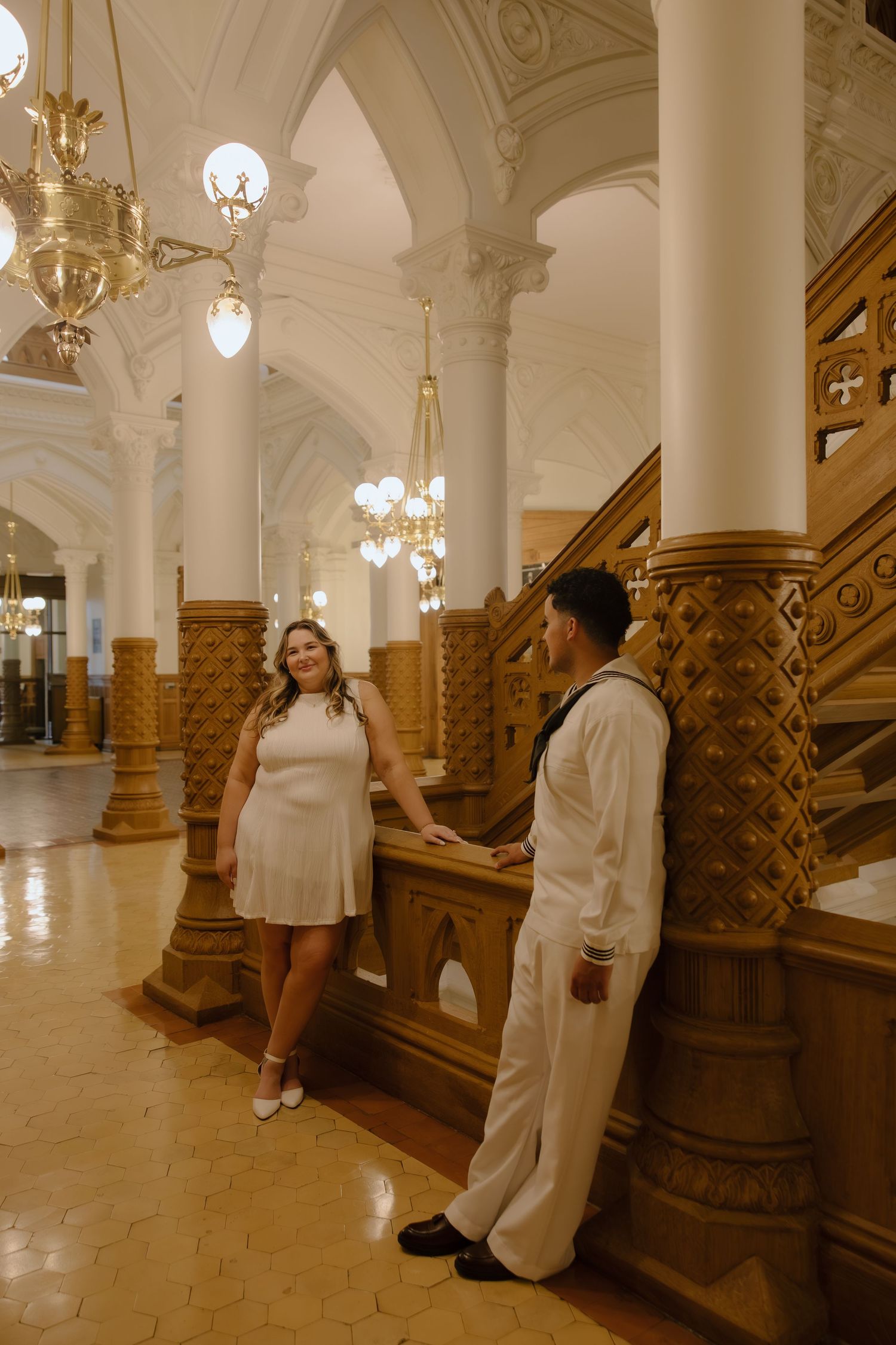 Couple in white attire poses by ornate wooden staircase in elegant hall with arched ceilings and chandeliers.