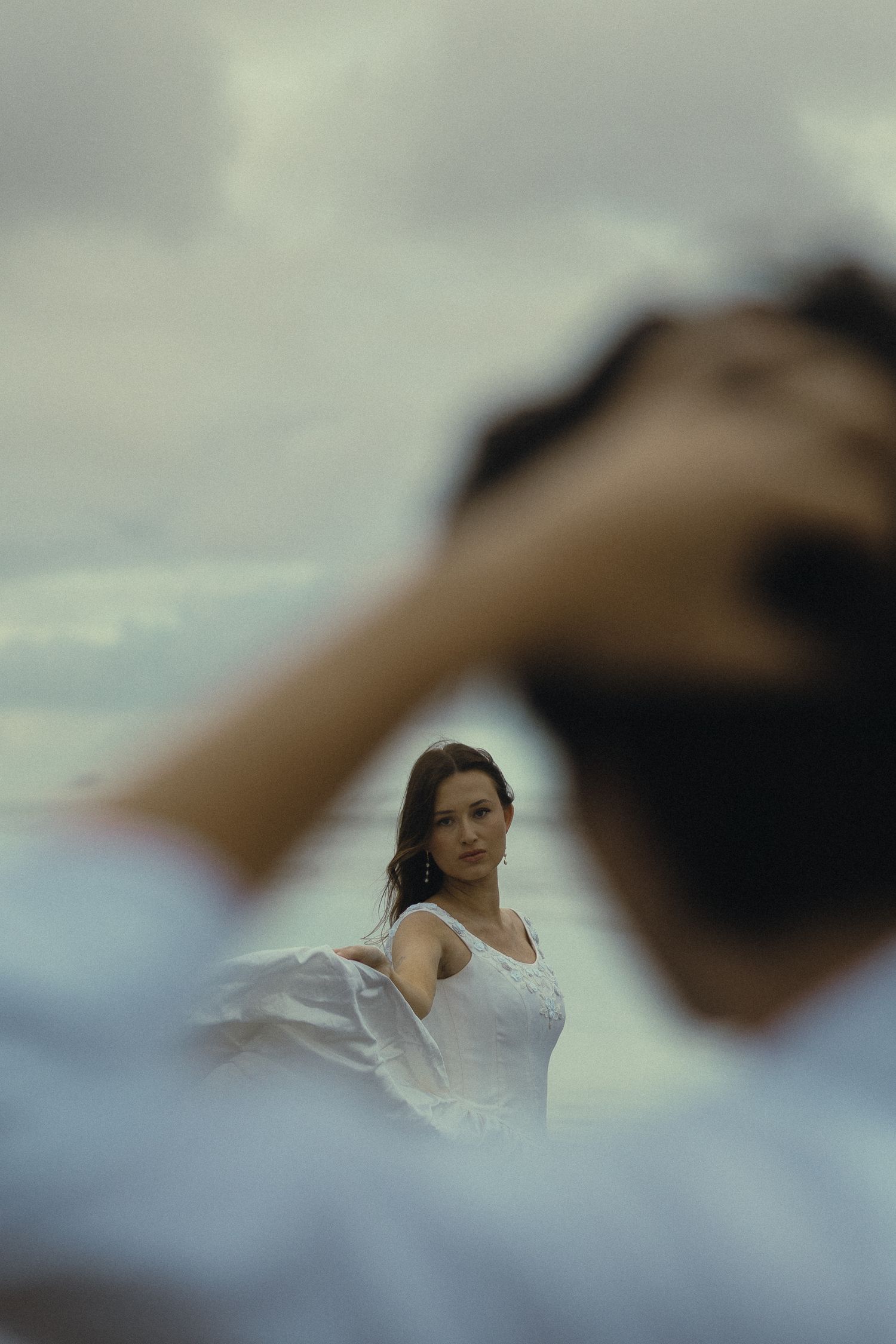 A woman in white dress stands in background as blurred man holds his head in foreground outdoors.
