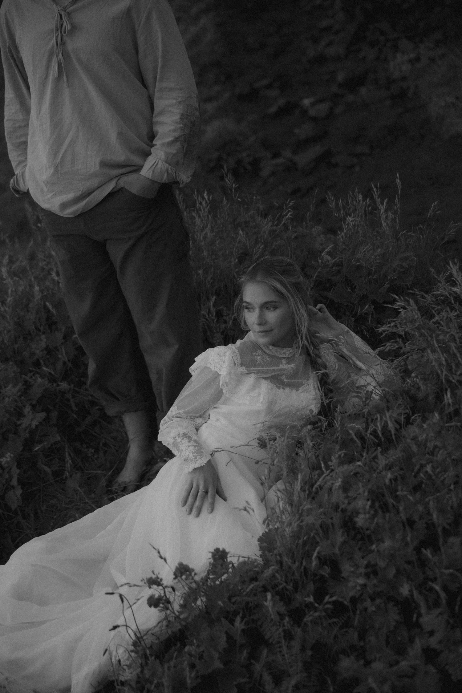Black and white photo of woman in white lace dress resting in garden while man stands beside her.