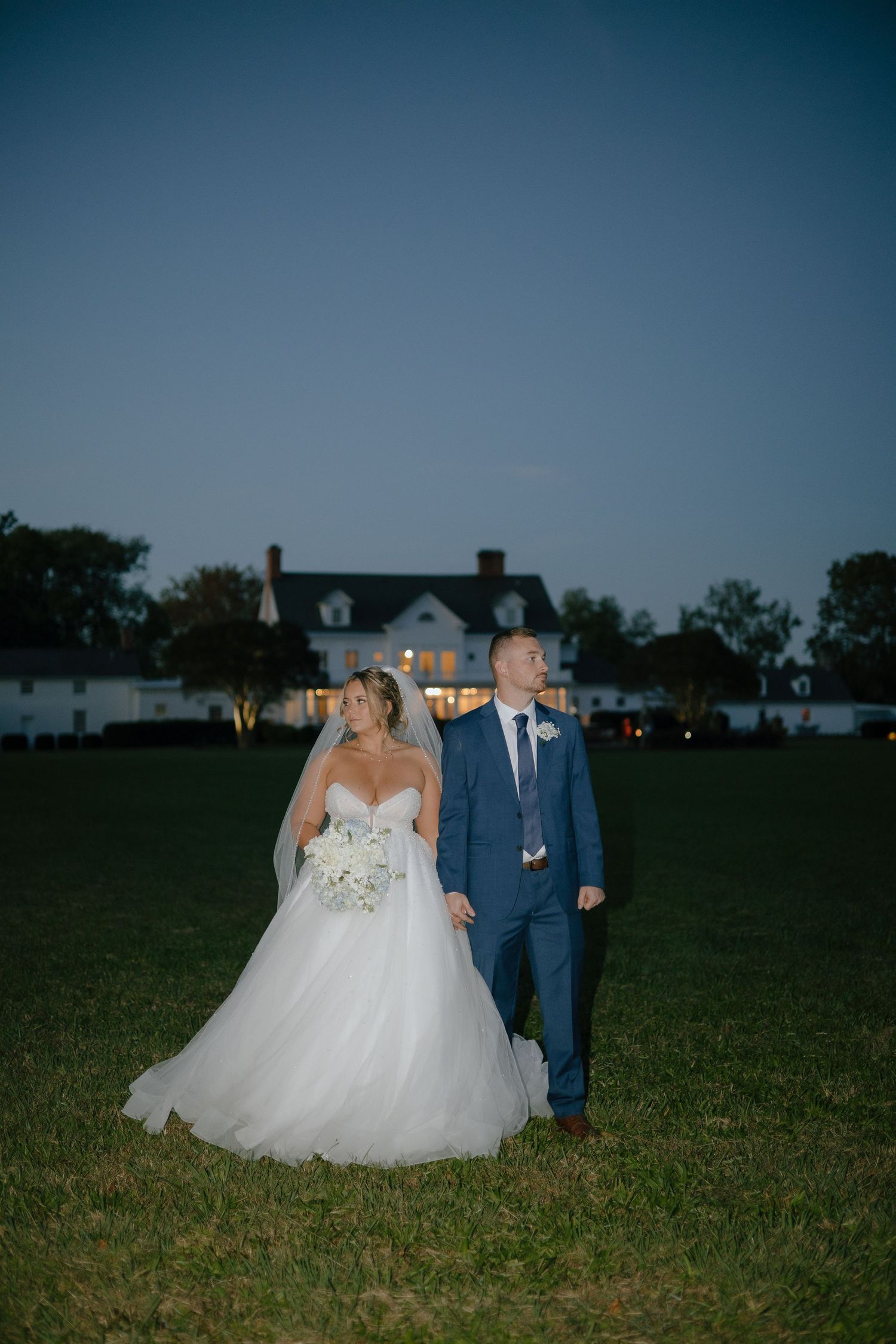 A couple in wedding attire walks through a grassy field at dusk with buildings in the background.
