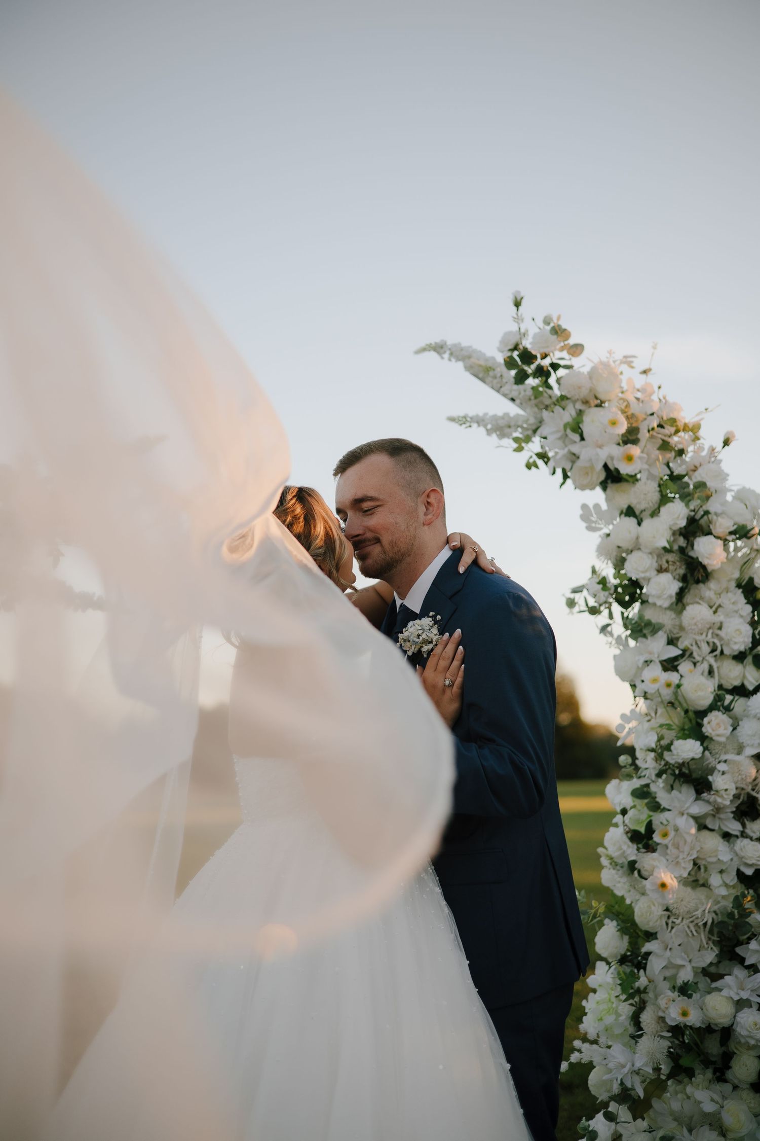 Wedding ceremony moment under a white floral arch with flowing veil in soft light.