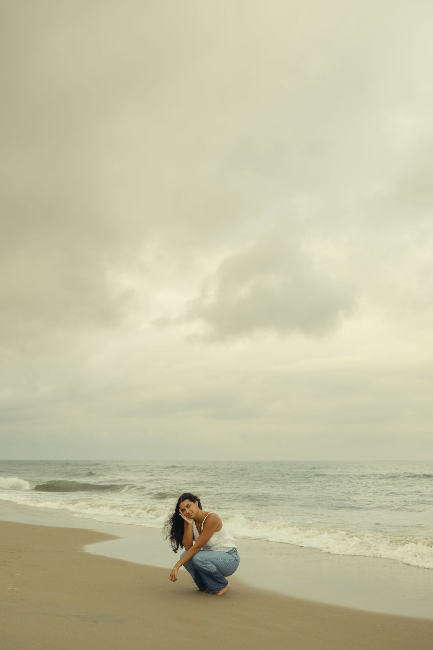 Woman crouching on a sandy beach near ocean waves under a cloudy sky, wearing jeans and a white top.