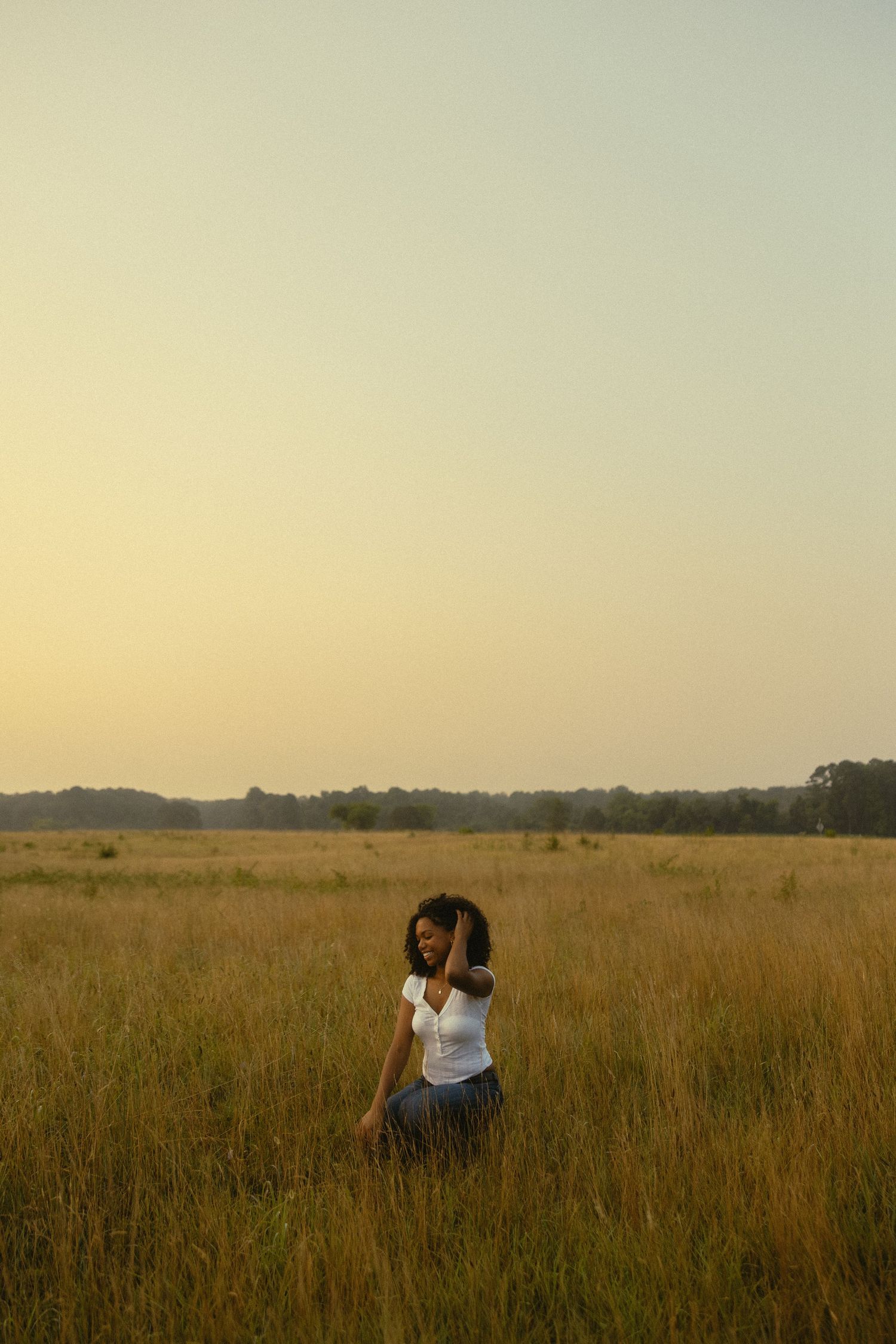 A young woman stands smiling in a golden field at sunset, wearing a white top and blue skirt.