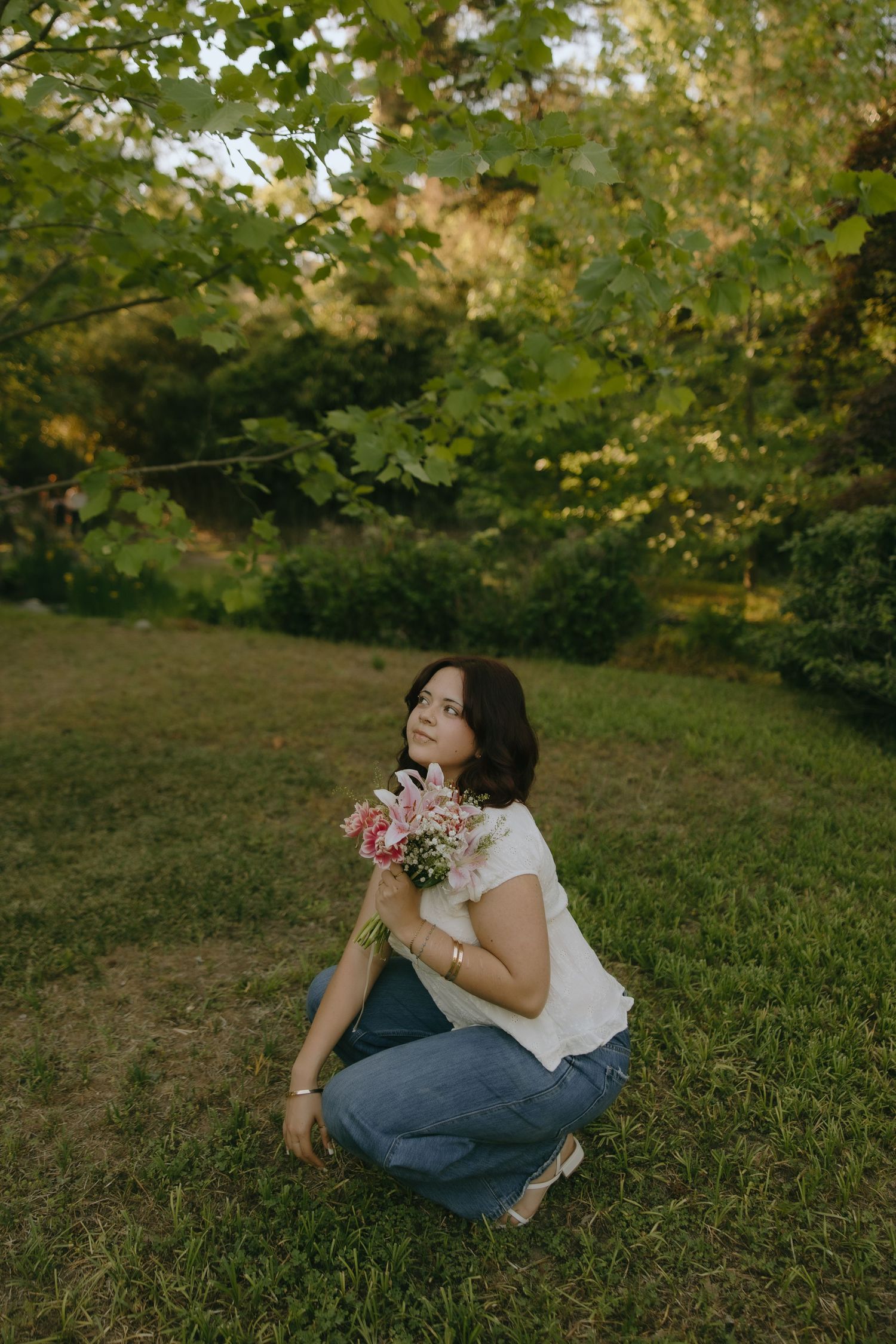 Woman crouching on grass holding pink flowers bouquet, wearing white top and jeans, surrounded by lush green trees.