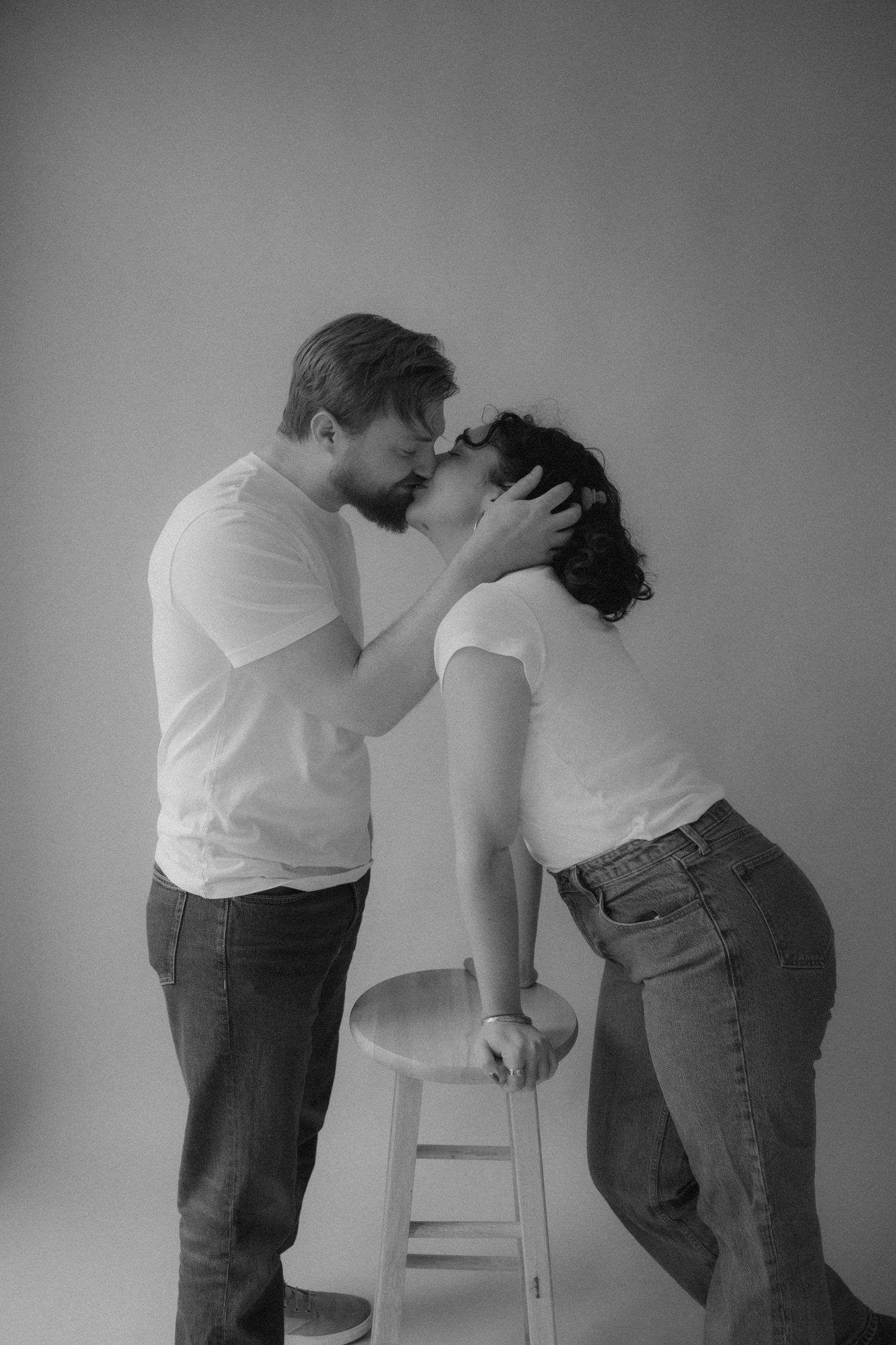 Black and white photo of a couple sharing a romantic kiss, both wearing white t-shirts and jeans in a studio setting.
