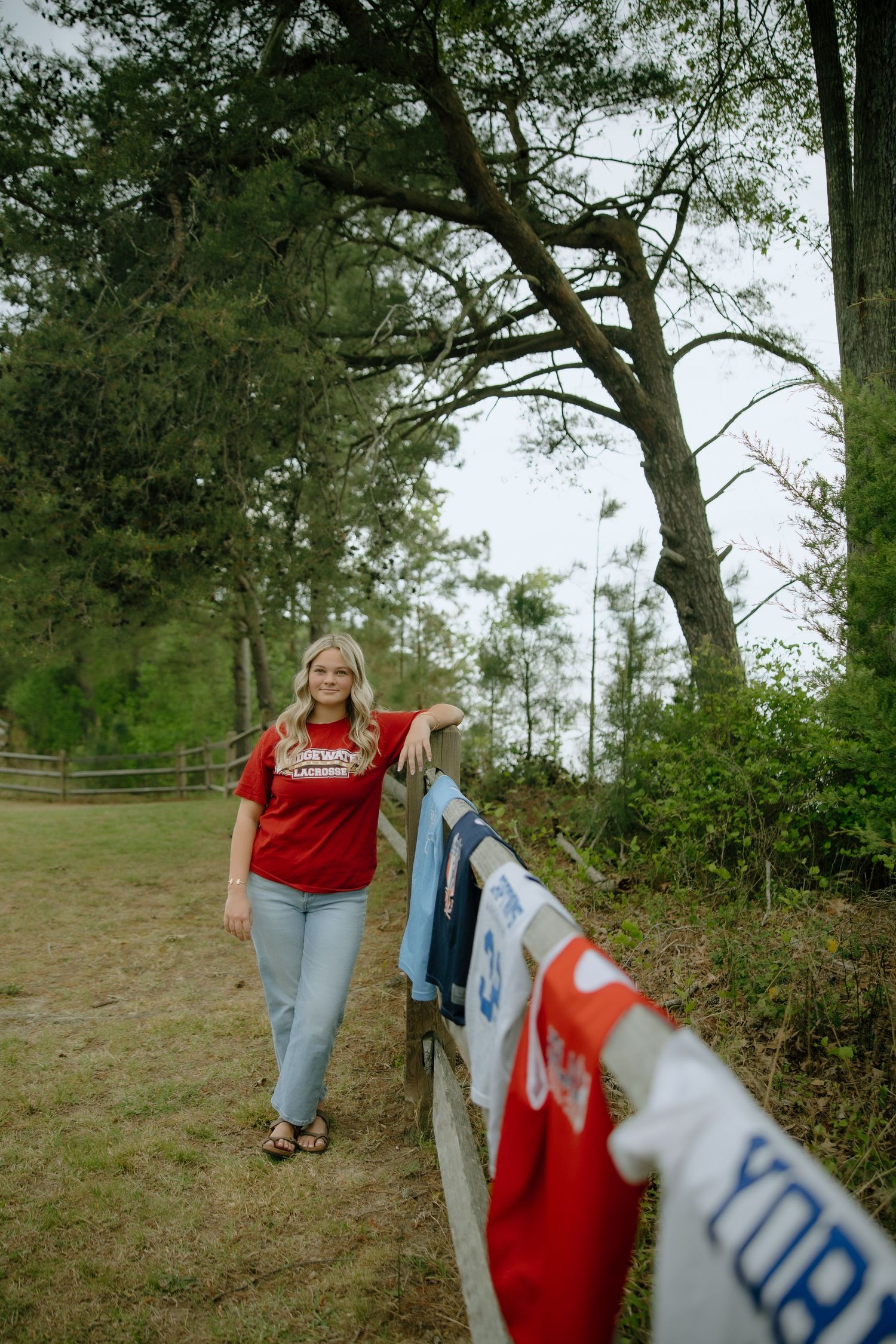 A young woman in a red t-shirt and jeans leans on a wooden fence with hanging shirts in a lush green outdoor setting.