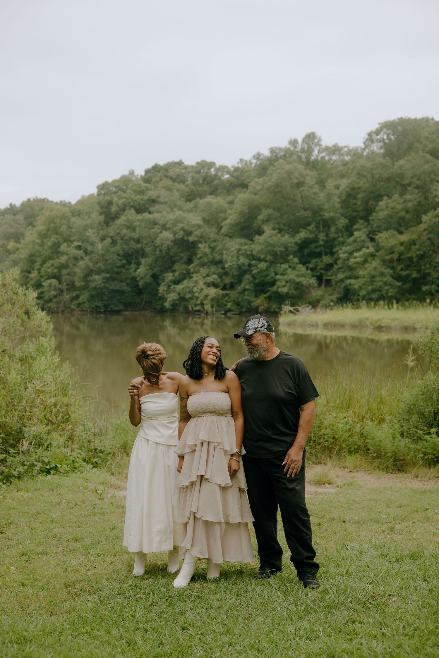Three people smiling and posing together near a serene pond surrounded by lush green trees on an overcast day.