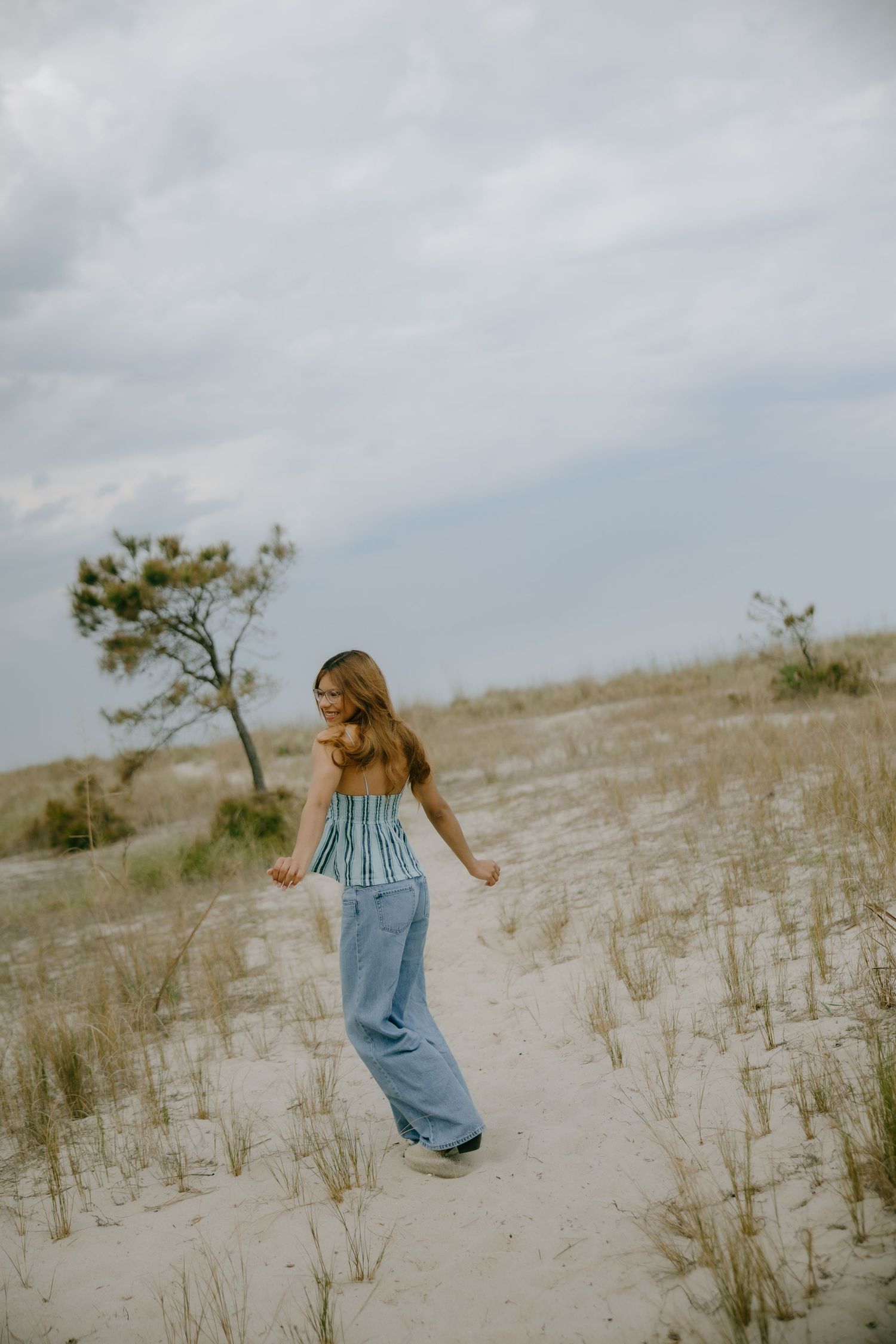 Young woman in blue wide-leg jeans and striped top walking on sandy dunes under cloudy sky.