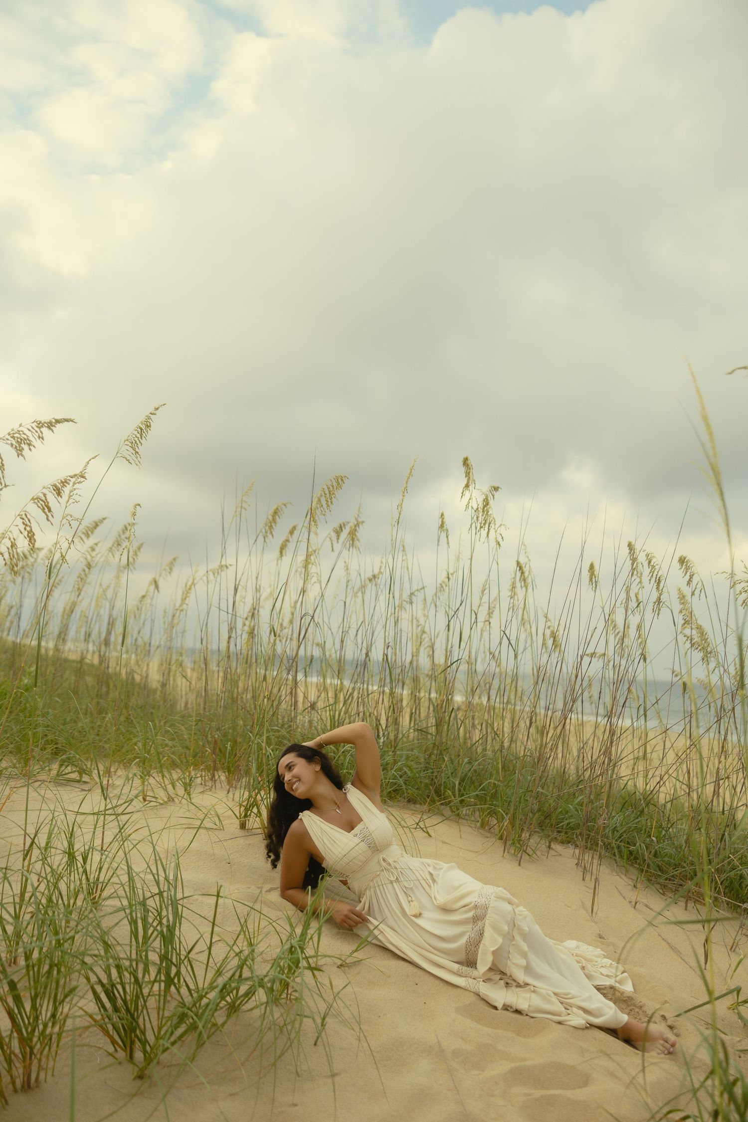 Woman in white dress reclining on sandy beach dunes with sea oats and cloudy sky backdrop.