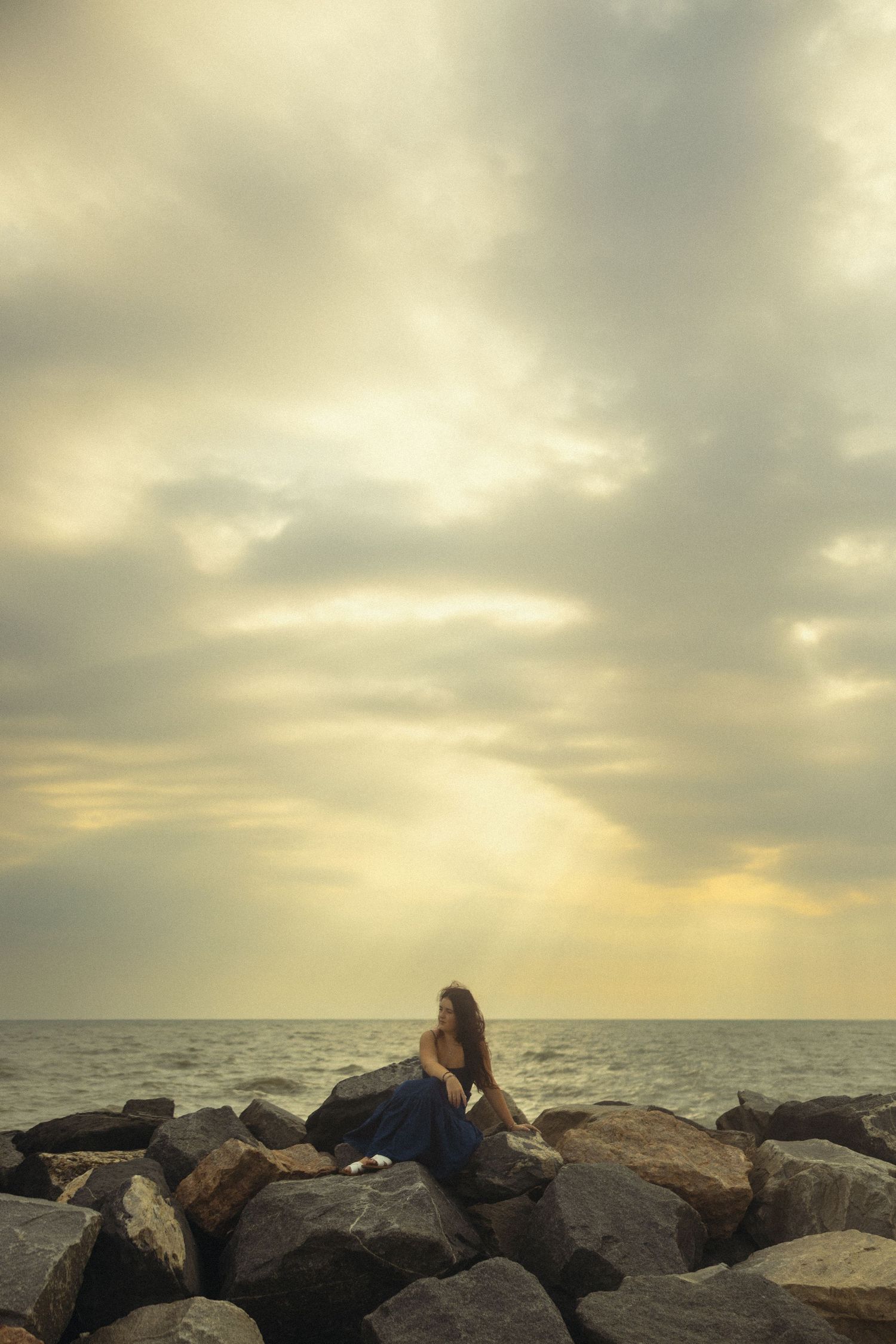 Woman in blue dress sitting on rocky shoreline at sunset with dramatic cloudy sky and calm ocean in background.