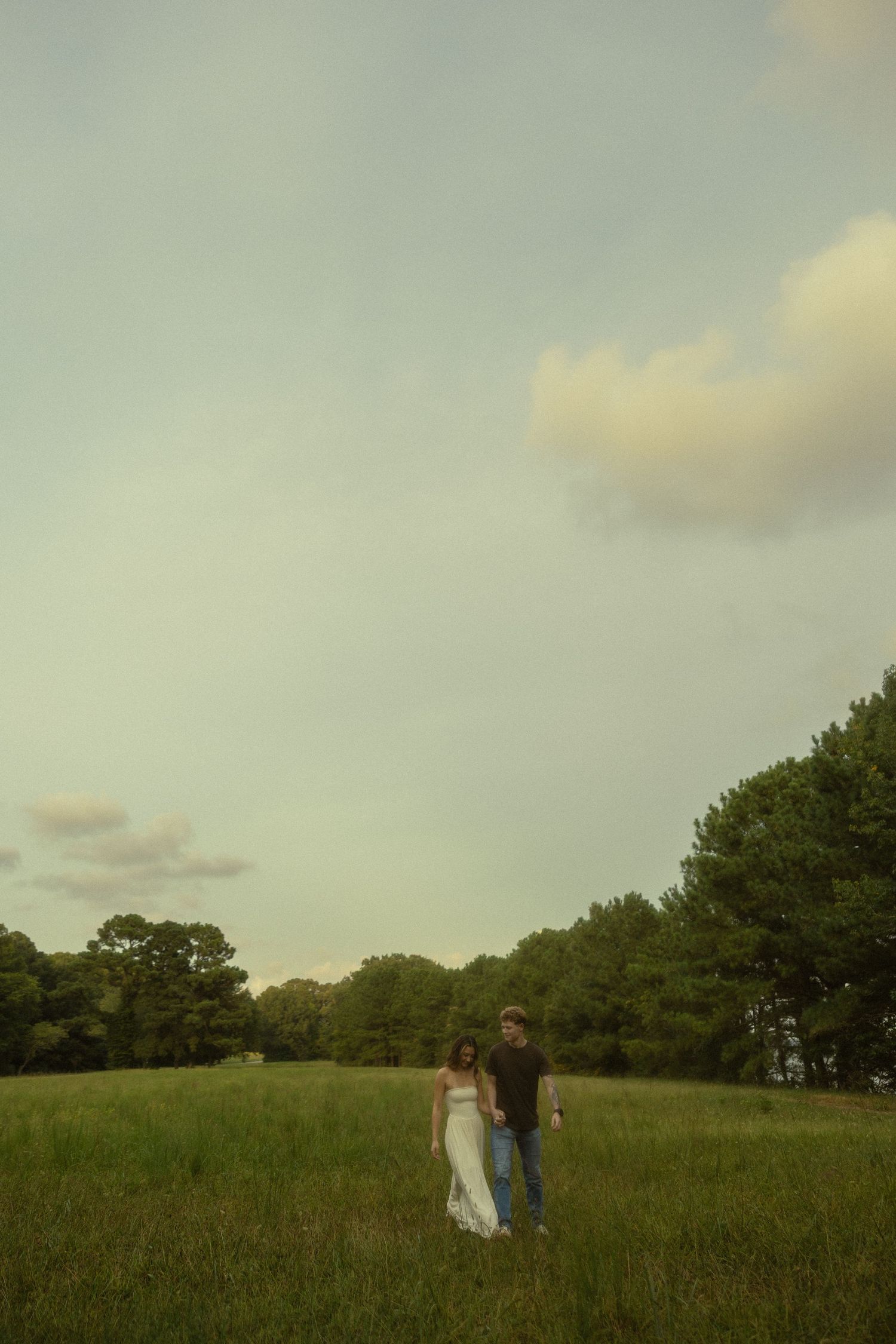 A couple stands in a lush green meadow surrounded by trees under a cloudy sky, woman in white dress.