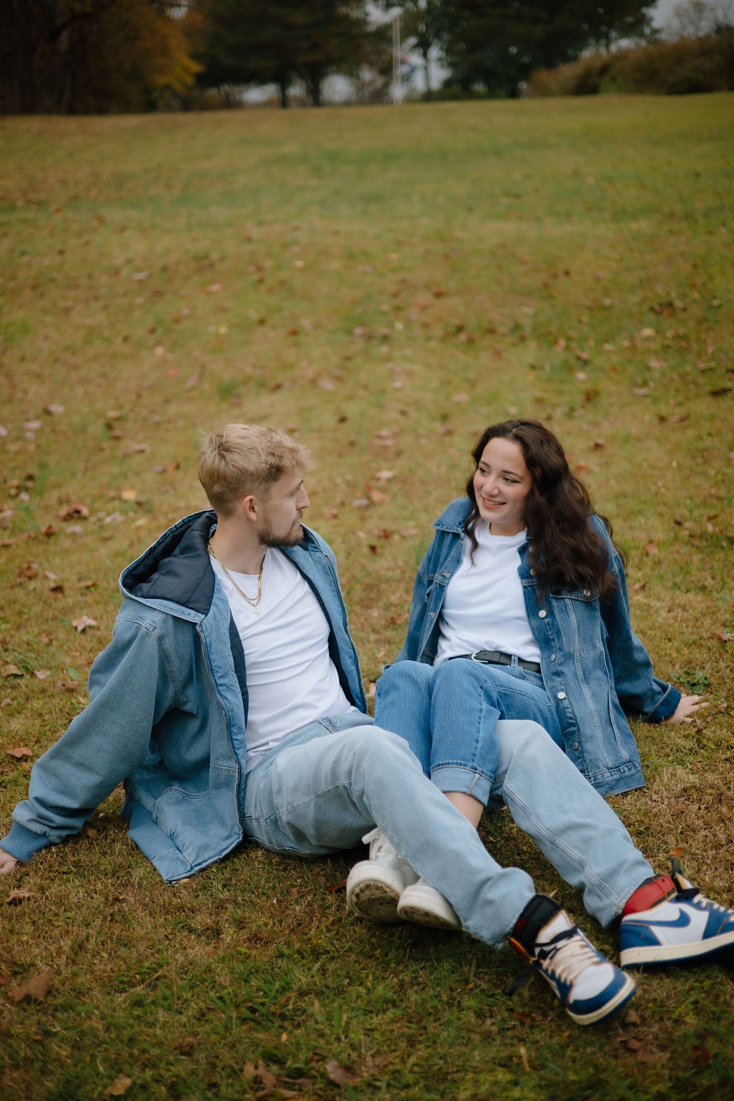 A couple relaxes on grass during autumn, wearing casual denim outfits and sneakers.