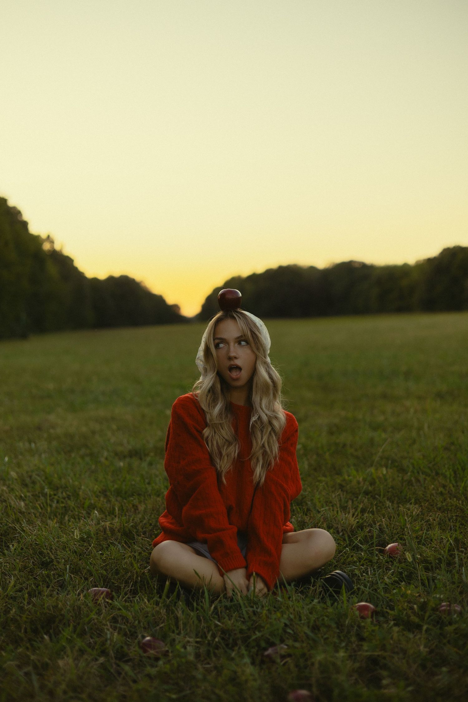 Blonde woman in red sweater sits cross-legged on grass at sunset with an apple balanced on her head.