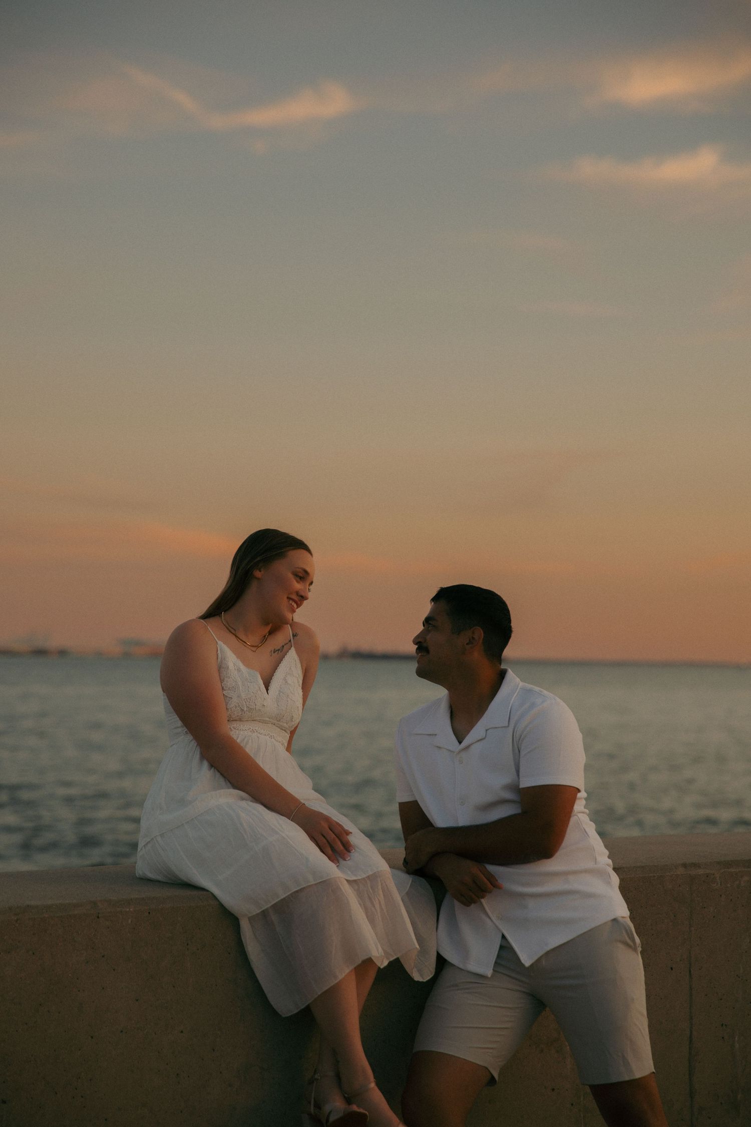 Couple sitting on a waterfront wall at sunset, smiling at each other, wearing white outfits with ocean in background.