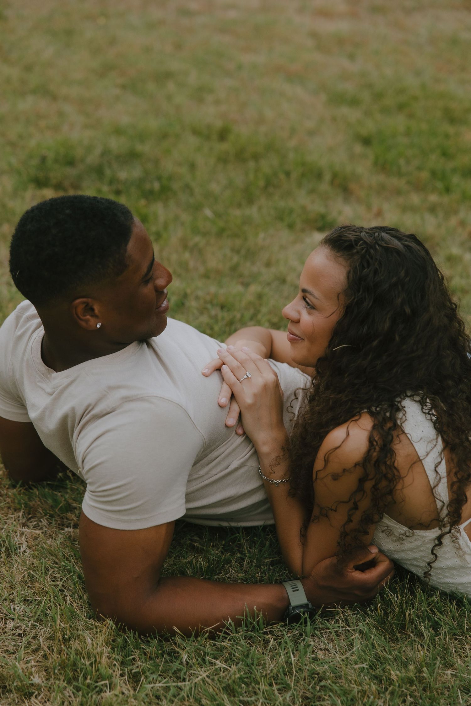 A couple lying on grass, holding hands and gazing at each other affectionately during an outdoor engagement session.
