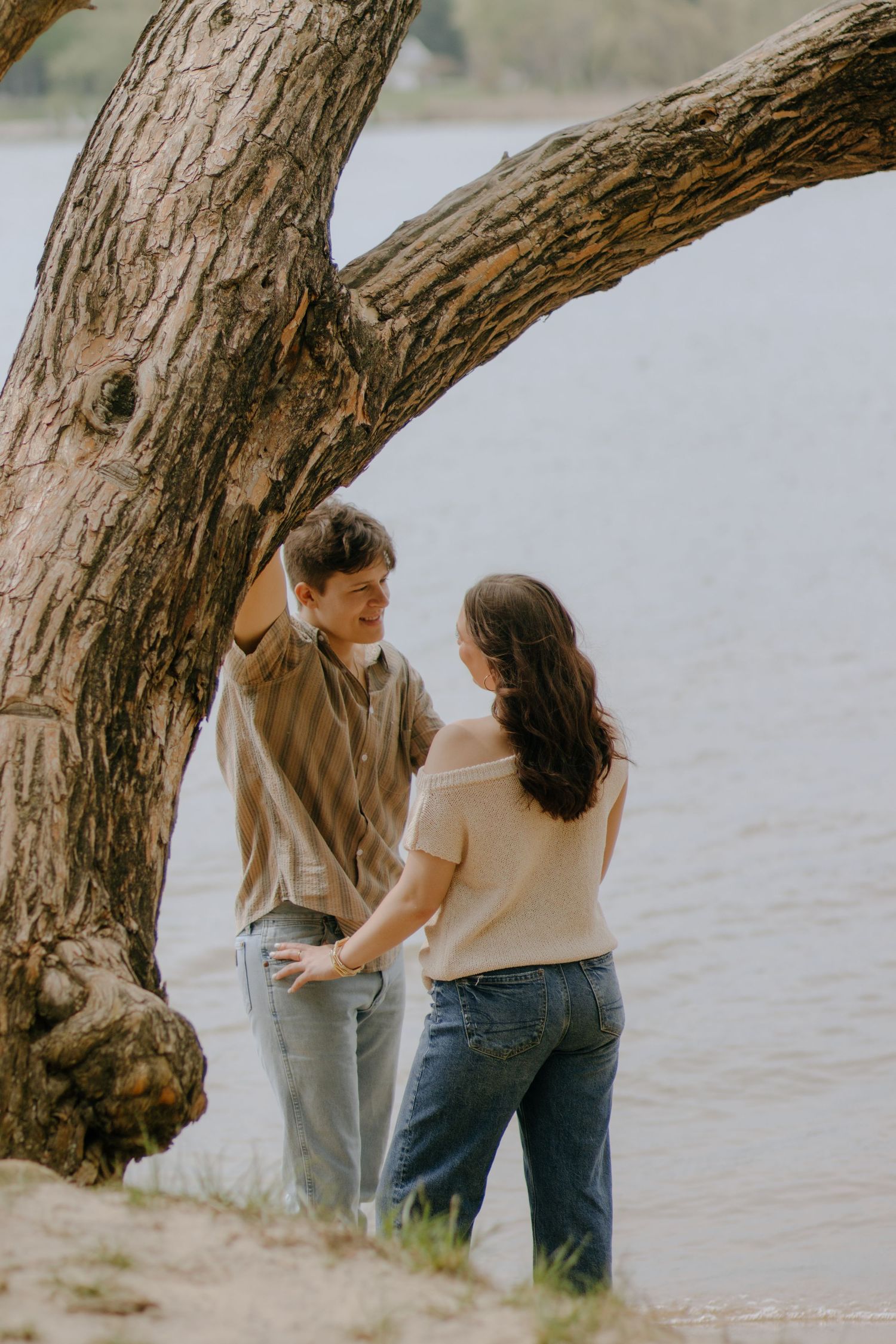 A couple shares intimate moments by a large tree trunk near the water.
