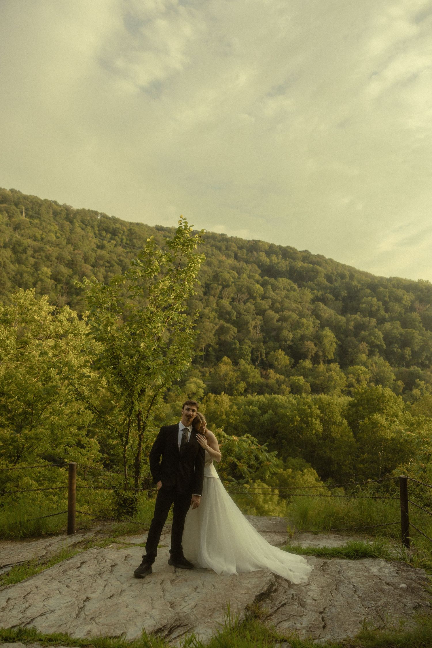 Bride and groom posing on rocky terrain with lush green forested hills and cloudy sky backdrop.