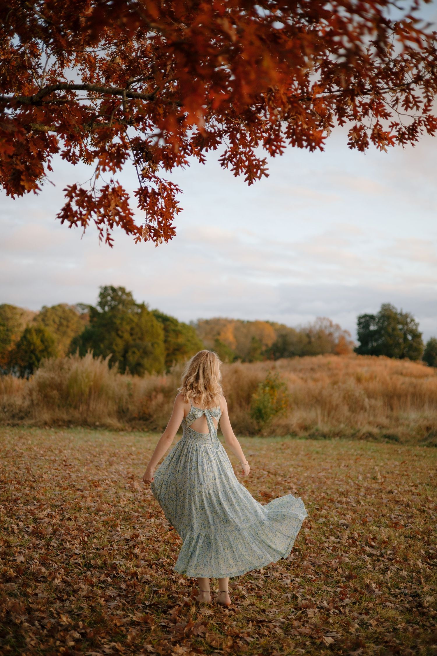 Senior in a flowing sage green dress twirls through autumn leaves beneath red-orange foliage overhead.