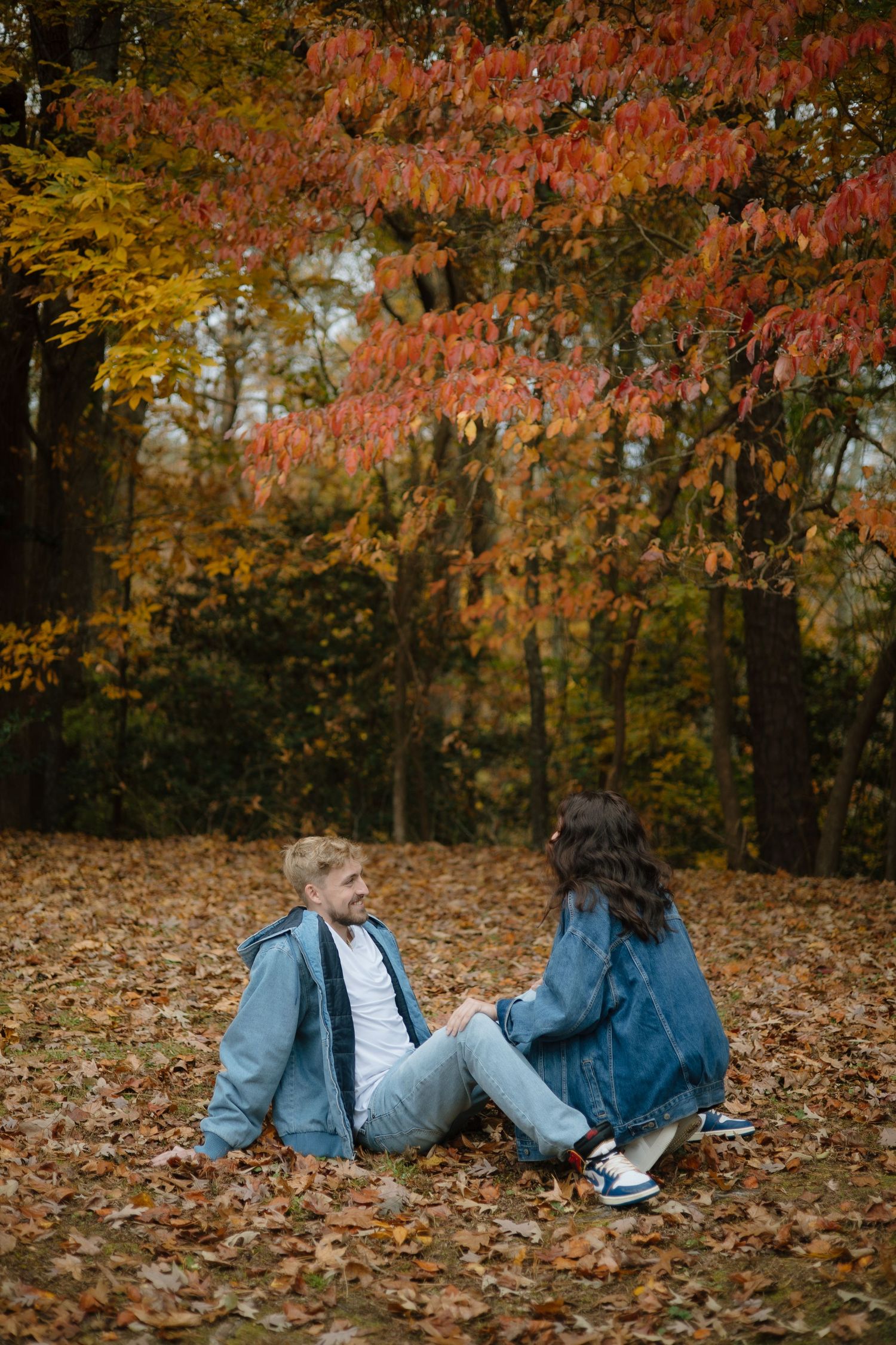 Two people in denim lying on autumn leaves having an intimate conversation in a park setting.