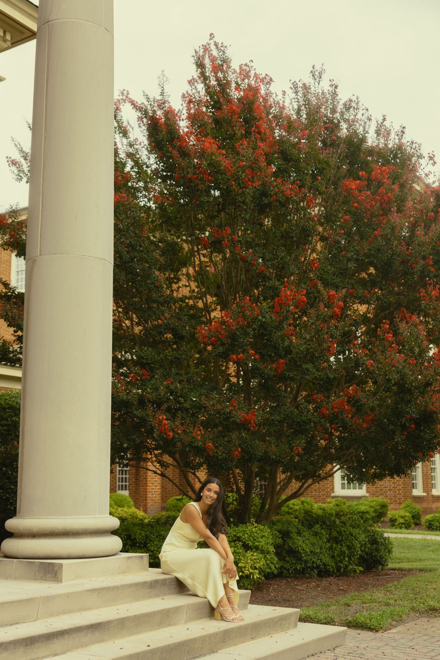 Woman in yellow skirt sits on steps by a white column, with a vibrant red flowering tree and brick building behind her.
